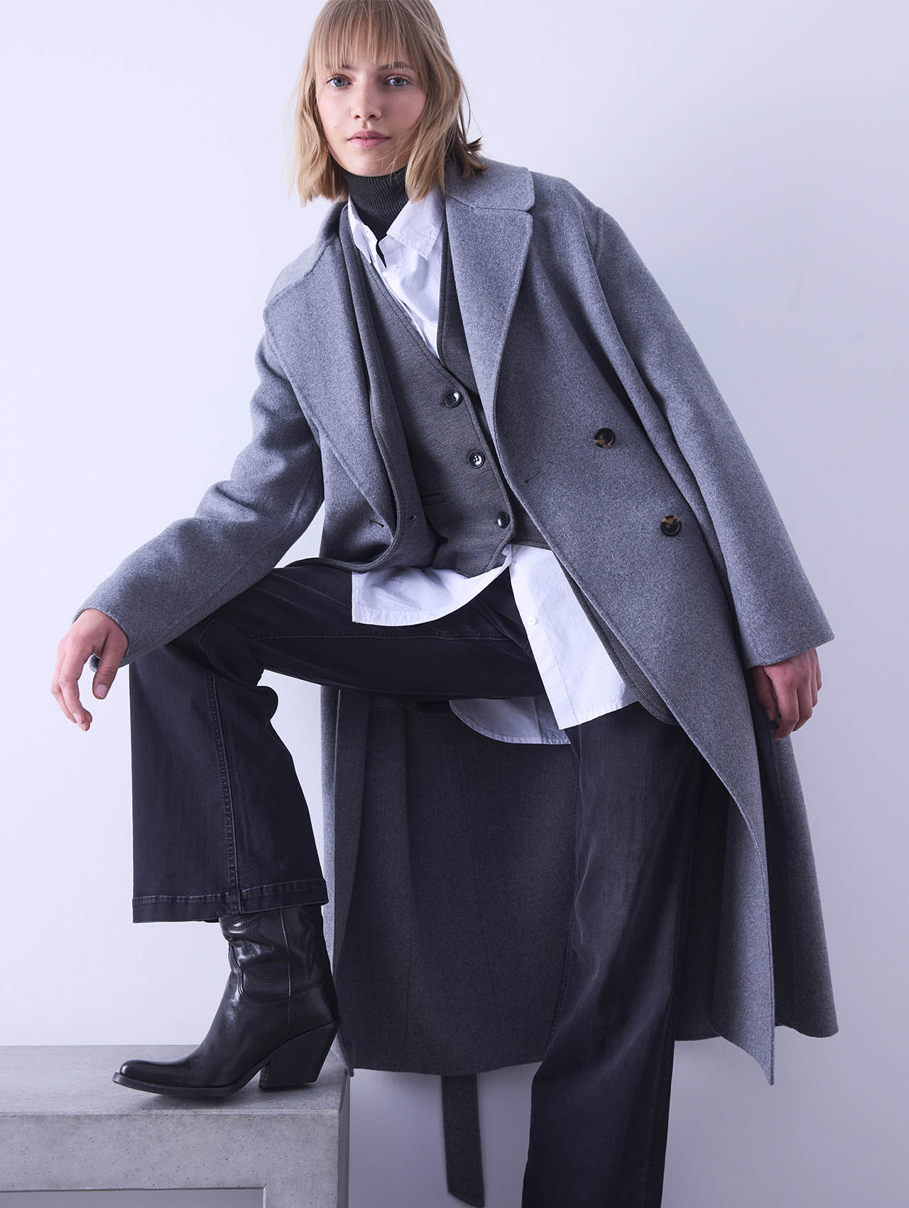 Eine Person mit blondem Haar trägt einen langen grauen Mantel, eine graue Weste, ein weißes Hemd, eine schwarze Hose und schwarze hochhackige Stiefel und stellt sich mit einem Fuß auf einen Sims vor einem hellen Hintergrund.