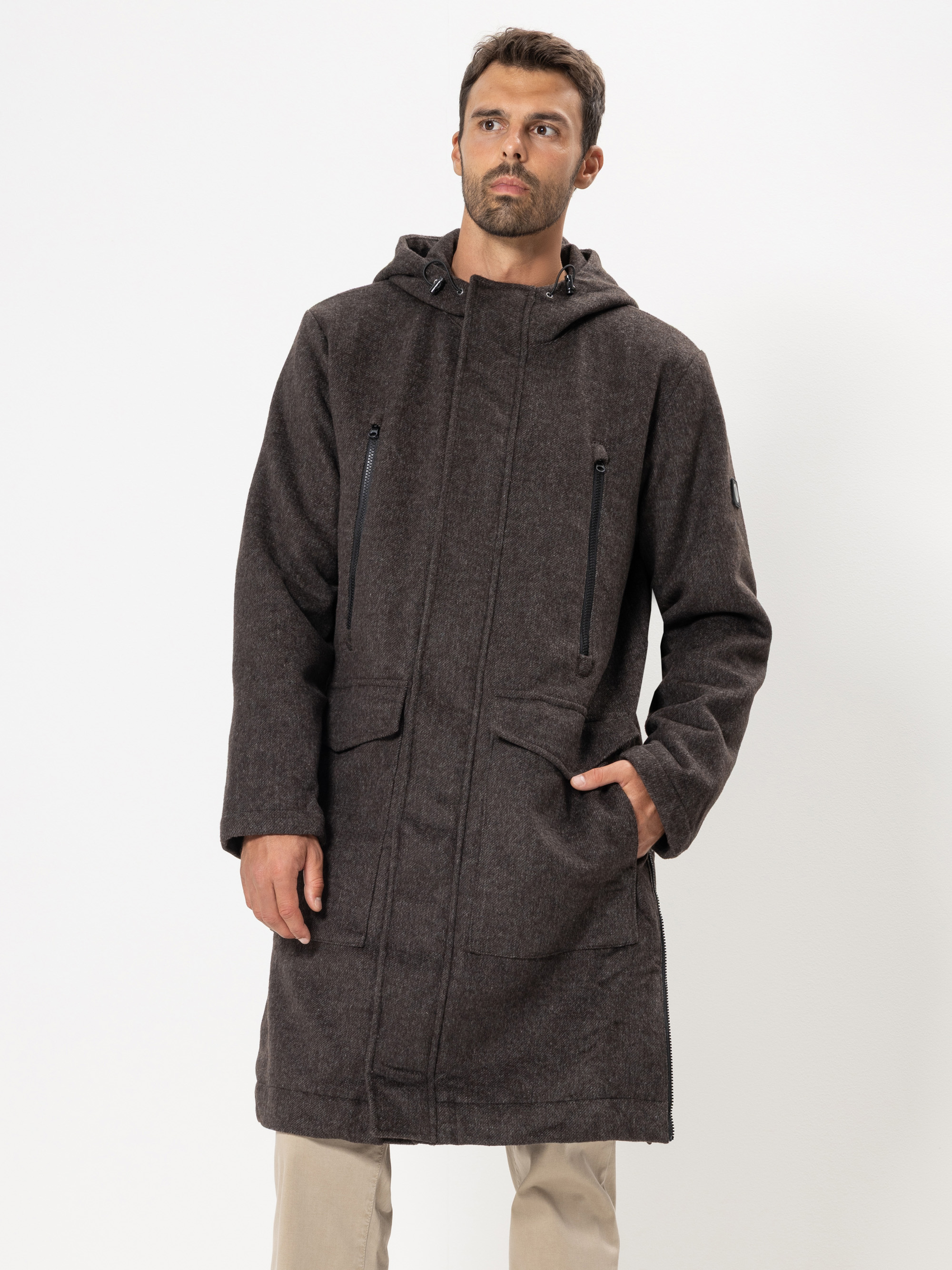 Ein Mann, der einen langen, dunkelbraunen Kapuzenmantel mit Reißverschlusstaschen trägt, steht vor einem schlichten weißen Hintergrund. Er hat eine Hand in der Tasche und blickt leicht zur Seite.