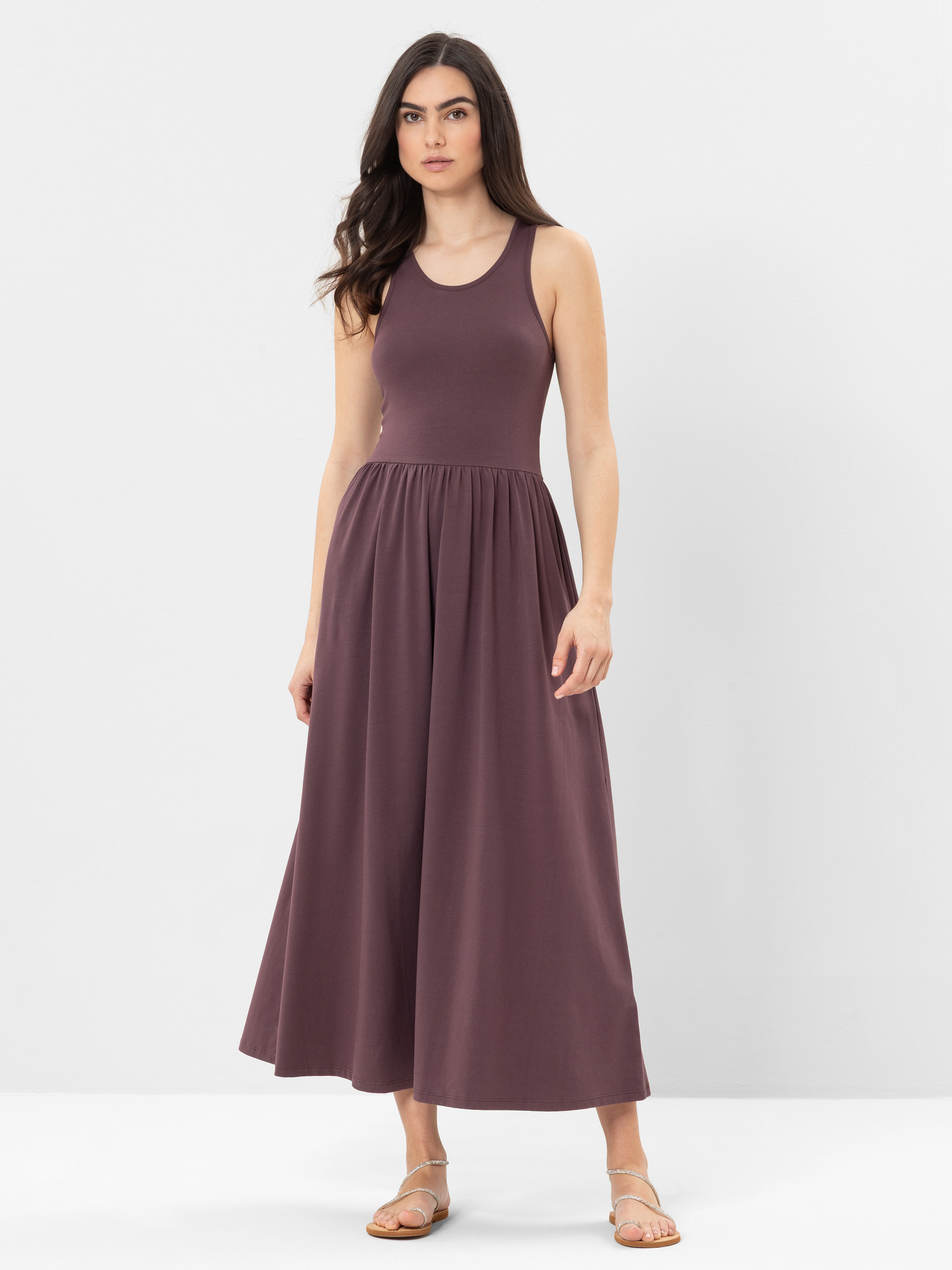 Eine Frau steht vor einem einfarbig weißen Hintergrund und trägt ein ärmelloses, knöchellanges, lilafarbenes Kleid mit tailliertem Oberteil und ausgestelltem Rock, dazu flache Sandalen.