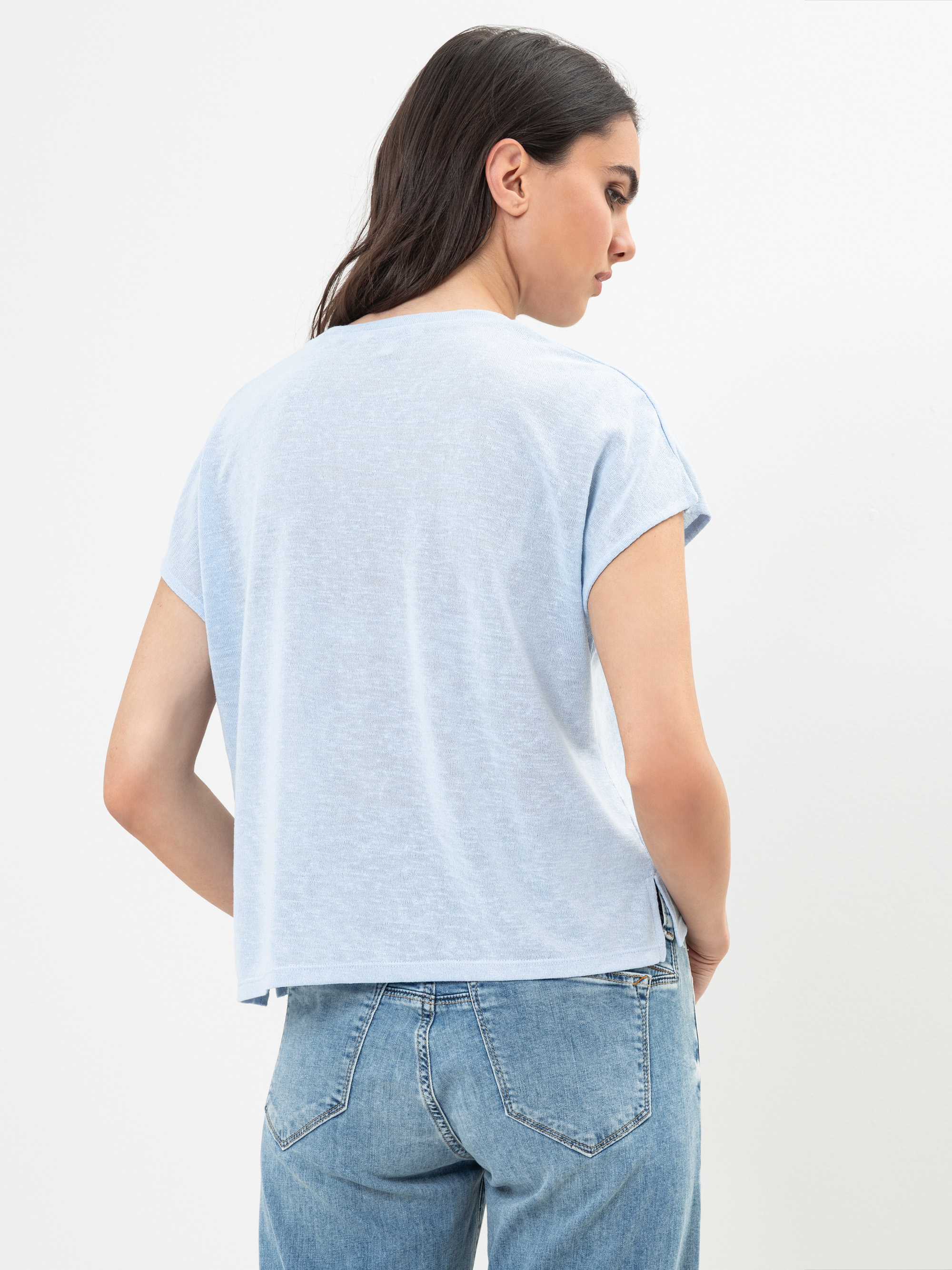 Eine Frau mit langen dunklen Haaren wird von hinten gezeigt. Sie trägt ein hellblaues, kurzärmeliges T-Shirt und blaue Jeans und steht vor einem schlichten weißen Hintergrund.