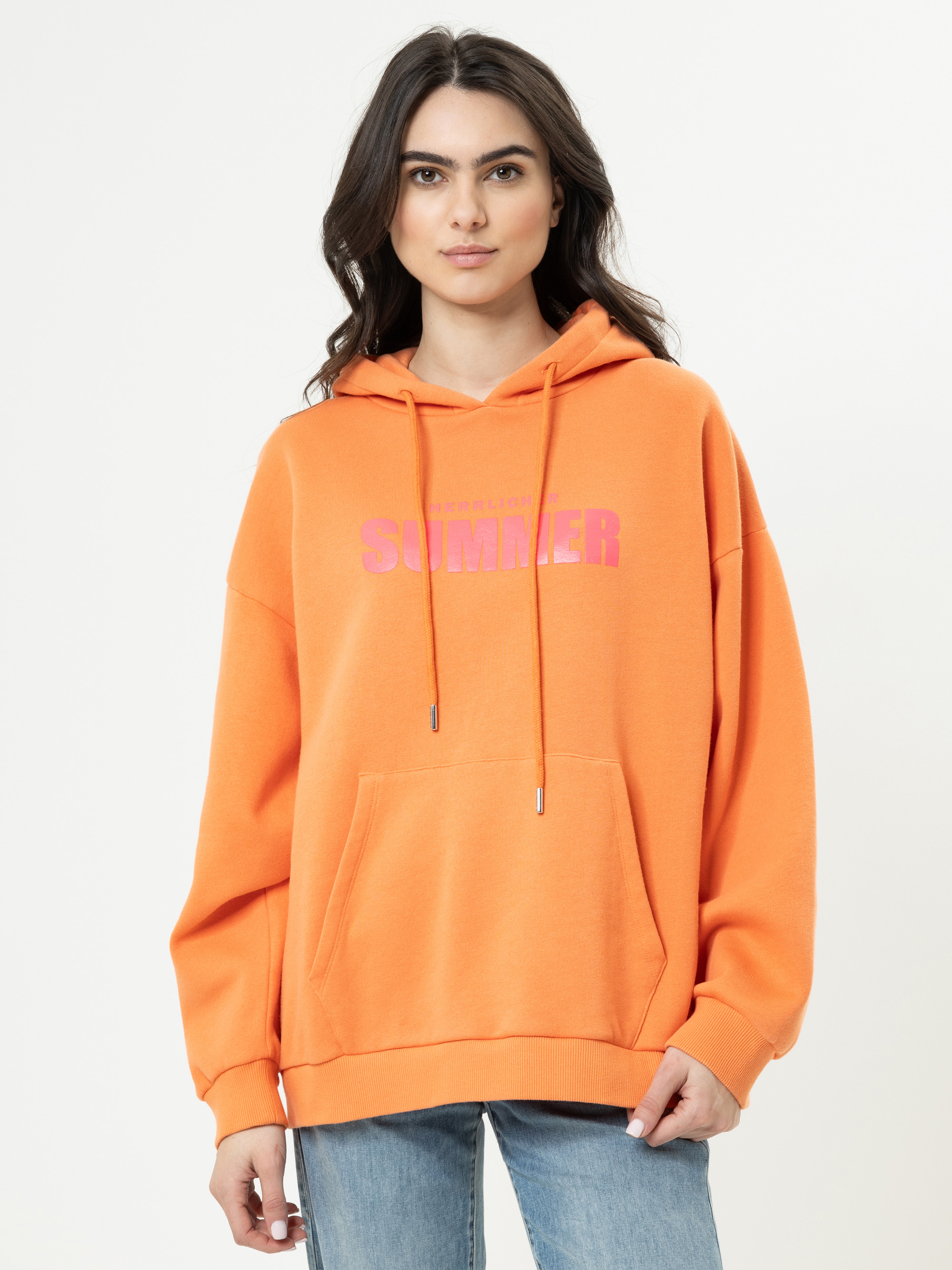Eine Frau mit langen dunklen Haaren trägt einen orangefarbenen Kapuzenpulli mit dem Wort SUMMER in Rosa, blaue Jeans und steht vor einem schlichten weißen Hintergrund.