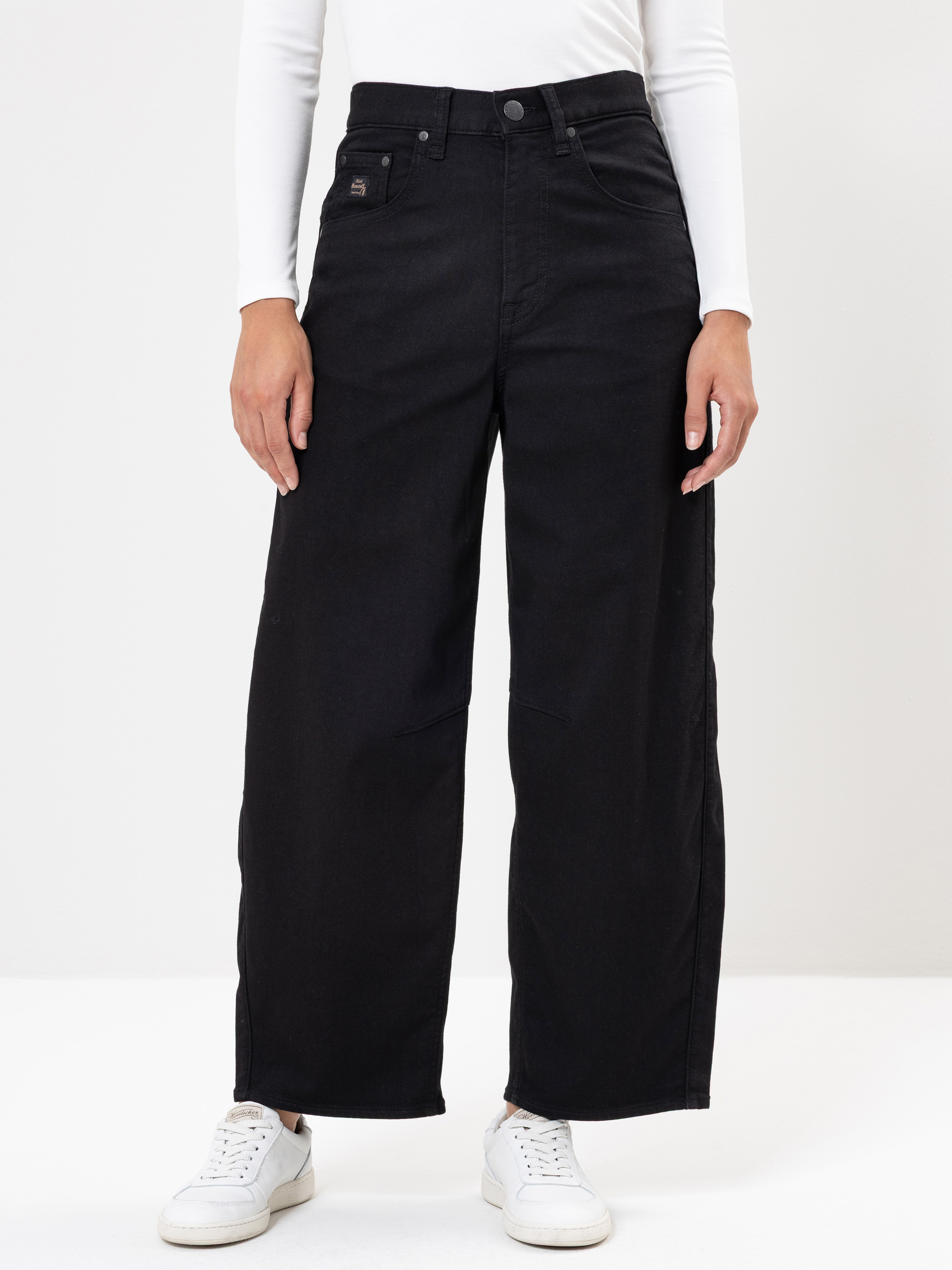 Eine Person, die eine schwarze Jeans mit hoher Taille und weitem Bein, ein weißes langärmeliges Oberteil und weiße Turnschuhe trägt, steht vor einem schlichten weißen Hintergrund. Die Hände der Person ruhen an der Seite.