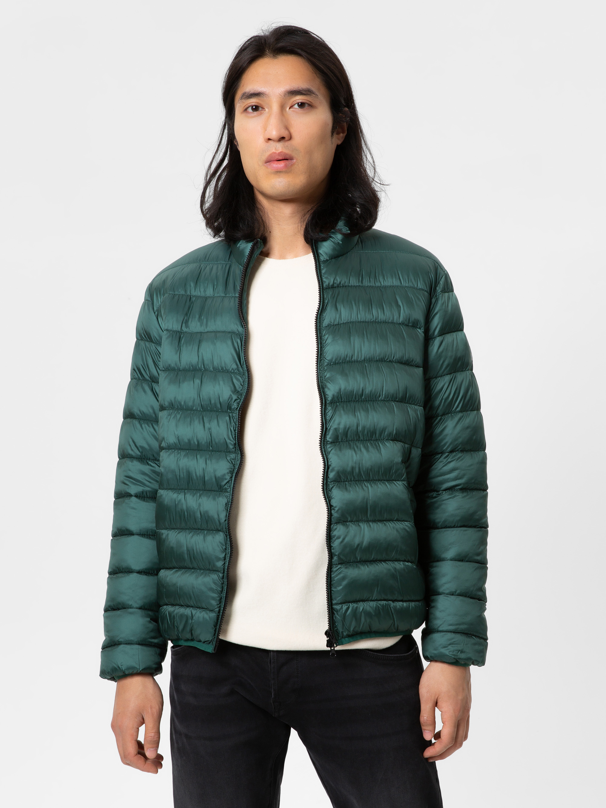 Eine Person mit langen dunklen Haaren trägt eine grüne Pufferjacke über einem weißen Hemd und einer schwarzen Hose und steht vor einem schlichten weißen Hintergrund.