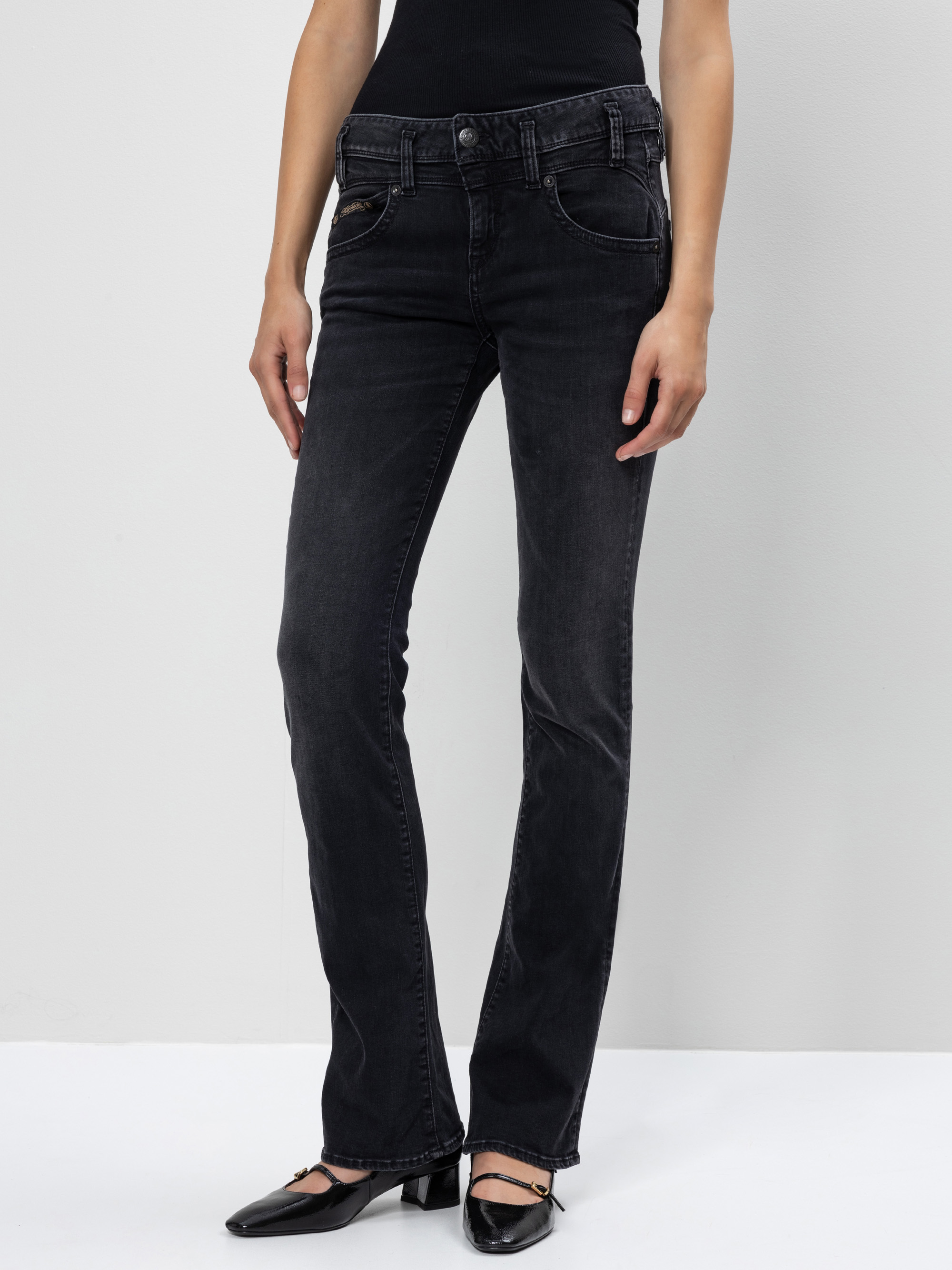 Eine Person in schwarzen Bootcut-Jeans und schwarzen Stöckelschuhen steht vor einem schlichten hellgrauen Hintergrund. Nur die untere Hälfte ihres Körpers ist sichtbar.