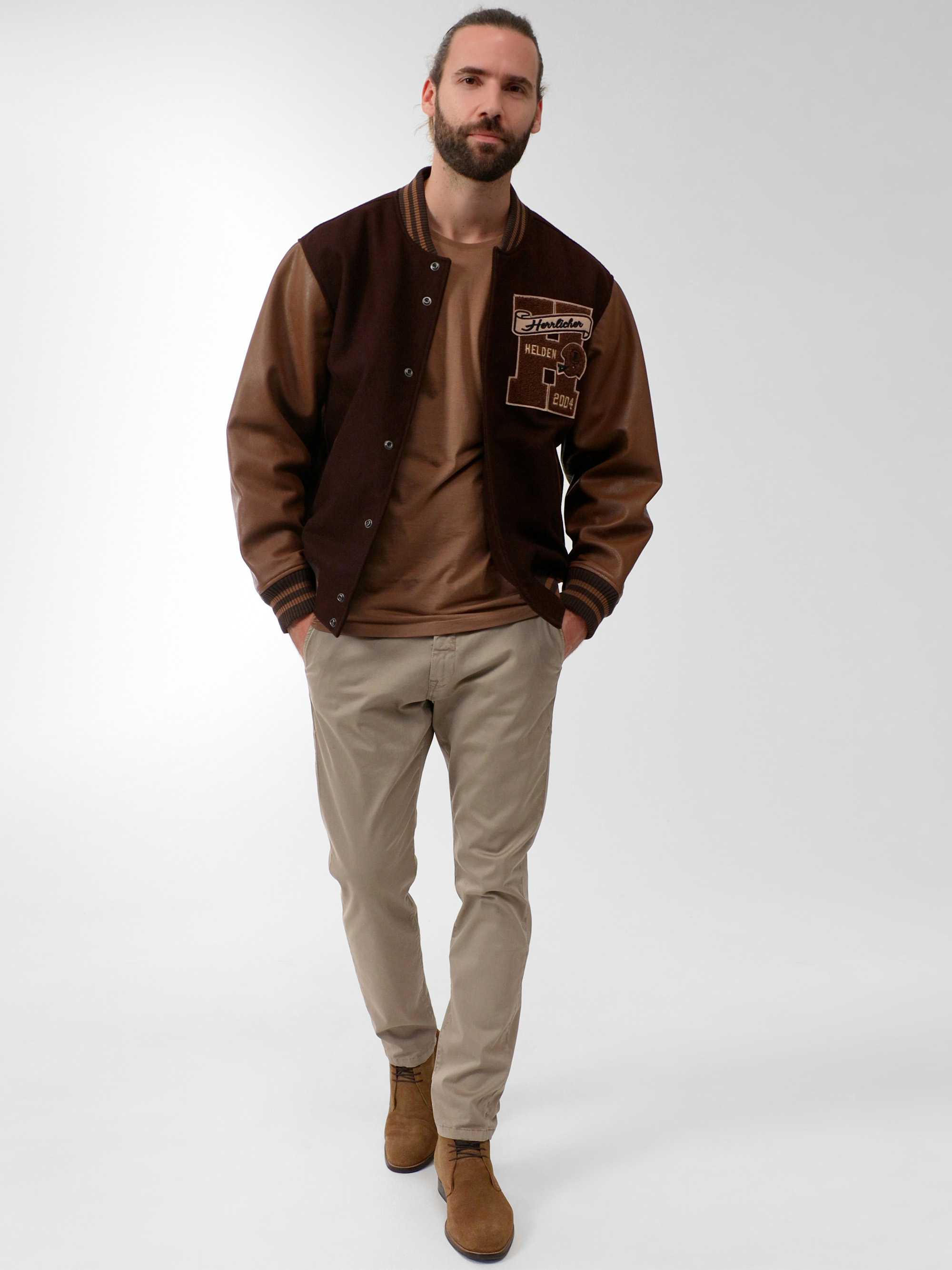 Ein Mann mit Bart trägt eine braune Sportjacke, ein beiges T-Shirt, eine helle khakifarbene Hose und braune Schuhe und geht selbstbewusst vor einem schlichten, hellen Hintergrund.