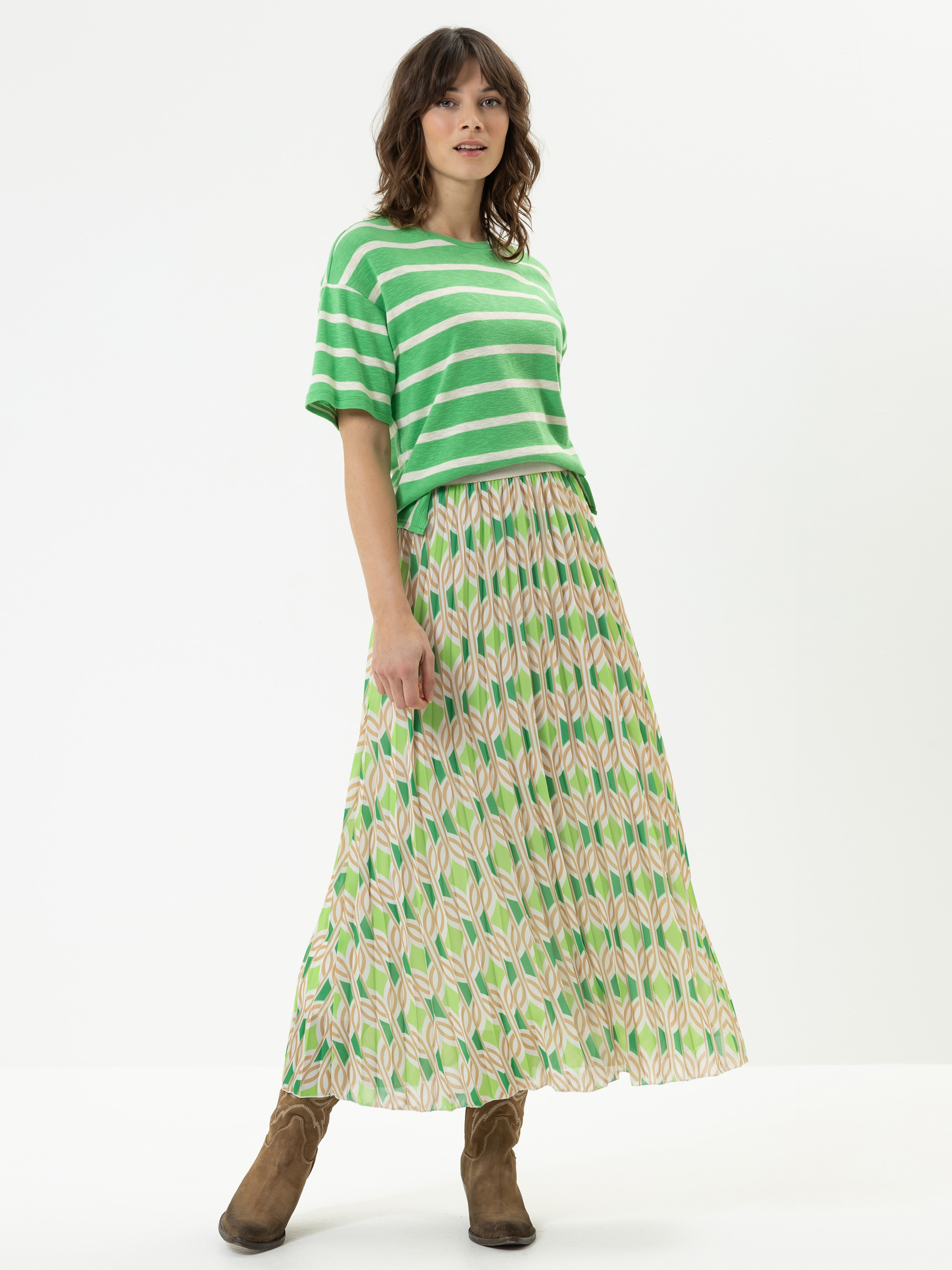 Eine Frau steht vor einem weißen Hintergrund und trägt ein grün-weiß gestreiftes, kurzärmeliges Oberteil, einen langen gemusterten Rock mit grünen und rosa Blattmustern und braune Stiefeletten. Sie hat gewelltes Haar und einen entspannten Gesichtsau