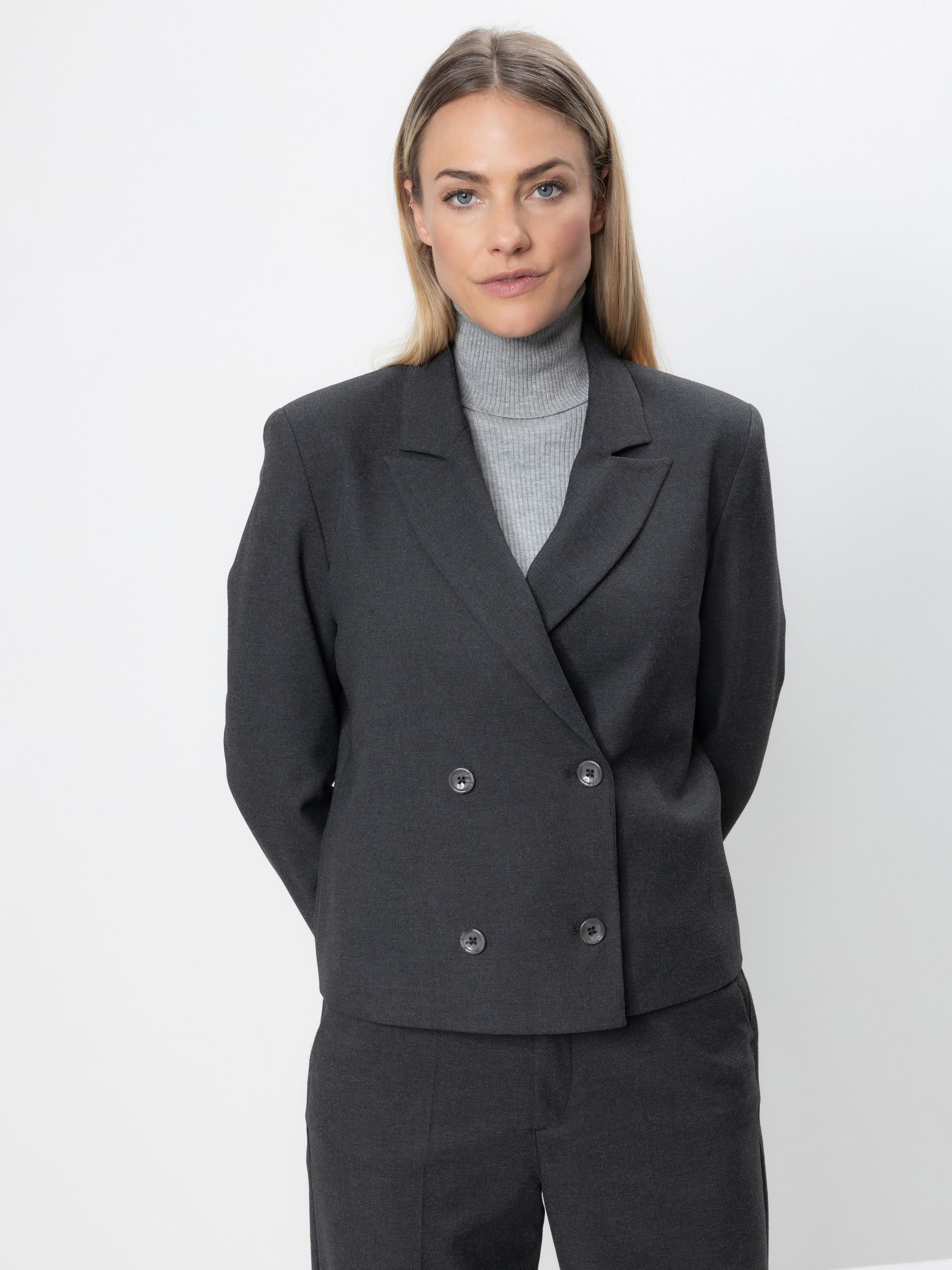 Eine Frau mit glattem, hellbraunem Haar trägt eine graue zweireihige Anzugsjacke und eine passende Hose über einem hellgrauen Rollkragenpullover. Sie steht vor einem schlichten weißen Hintergrund und blickt mit neutraler Miene nach vorn.