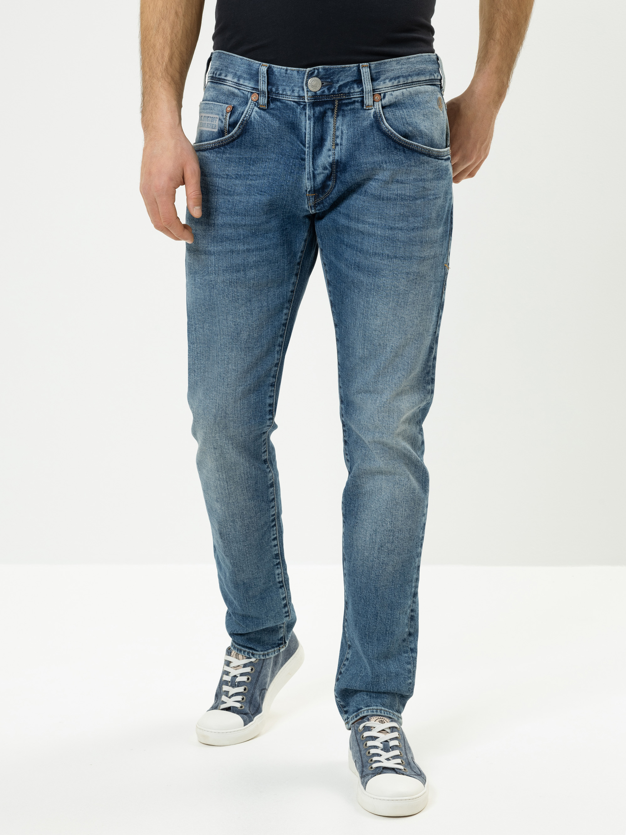 Eine Person in hellblauen Jeans und blauen Turnschuhen steht vor einem schlichten weißen Hintergrund und hat die Hände entspannt an der Seite. Ihr Oberkörper ist teilweise außerhalb des Bildausschnitts.