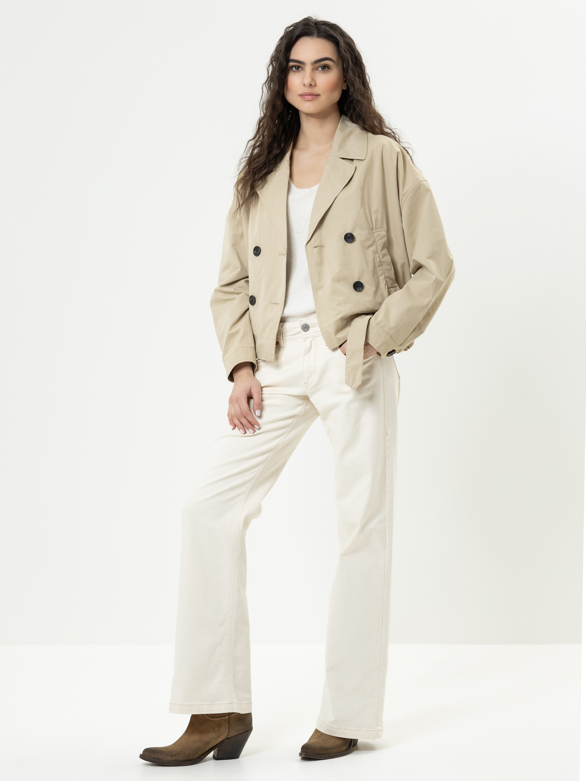 Eine Frau mit langem gewelltem Haar steht in neutraler Pose, trägt eine beigefarbene zweireihige Jacke, ein weißes Oberteil, eine weiße Hose mit weitem Bein und braune Stiefeletten vor einem schlichten weißen Hintergrund.