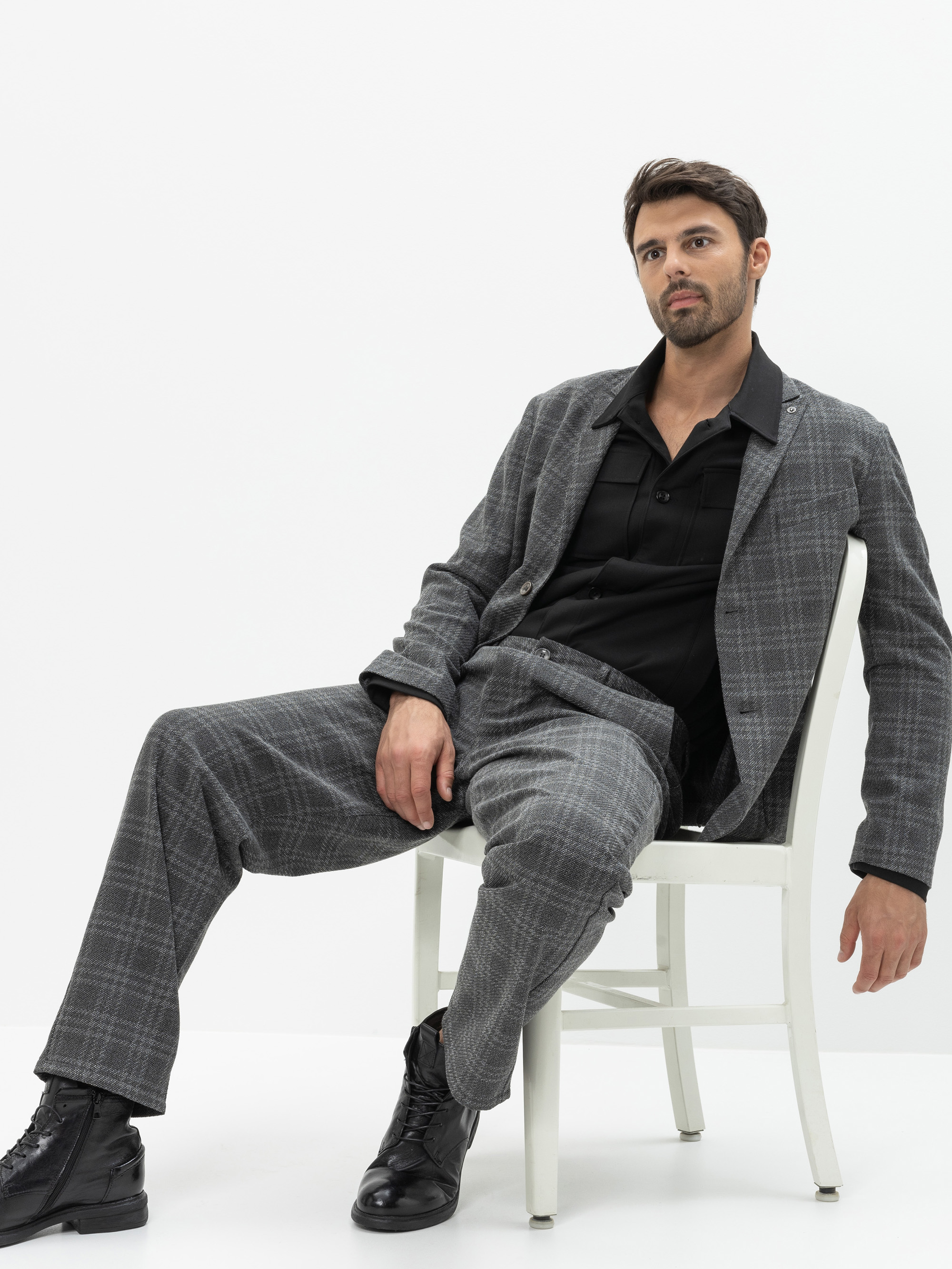 Ein Mann im grau-karierten Anzug und schwarzem Hemd sitzt lässig auf einem weißen Stuhl vor einem schlichten weißen Hintergrund, eine Hand ruht auf seinem Bein, die andere hängt vom Stuhl herunter.