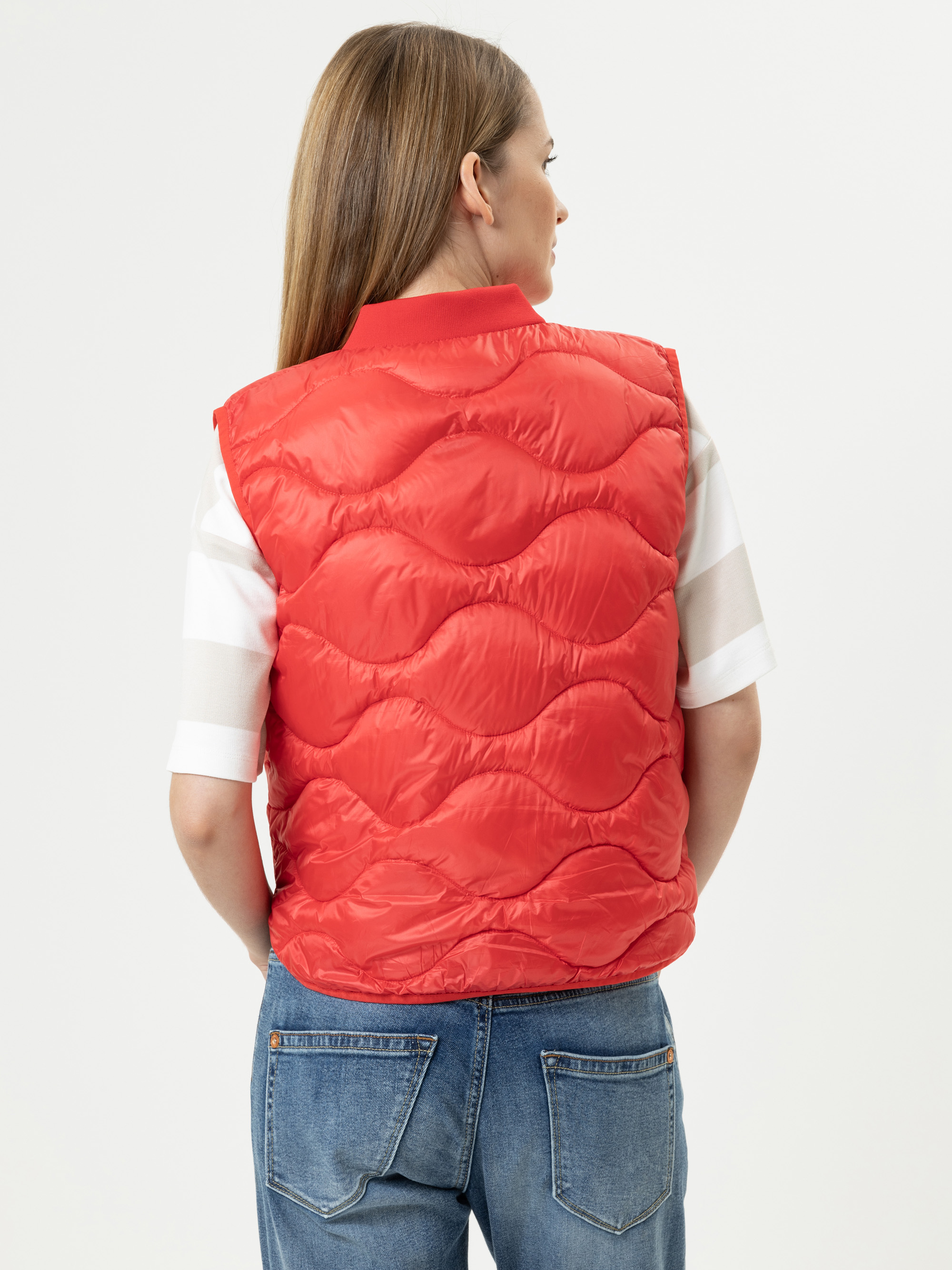 Eine Frau mit glattem, hellbraunem Haar ist von hinten zu sehen. Sie trägt eine leuchtend rote Steppweste über einem weißen kurzärmeligen Hemd und blaue Jeans und steht vor einem schlichten, hellen Hintergrund.