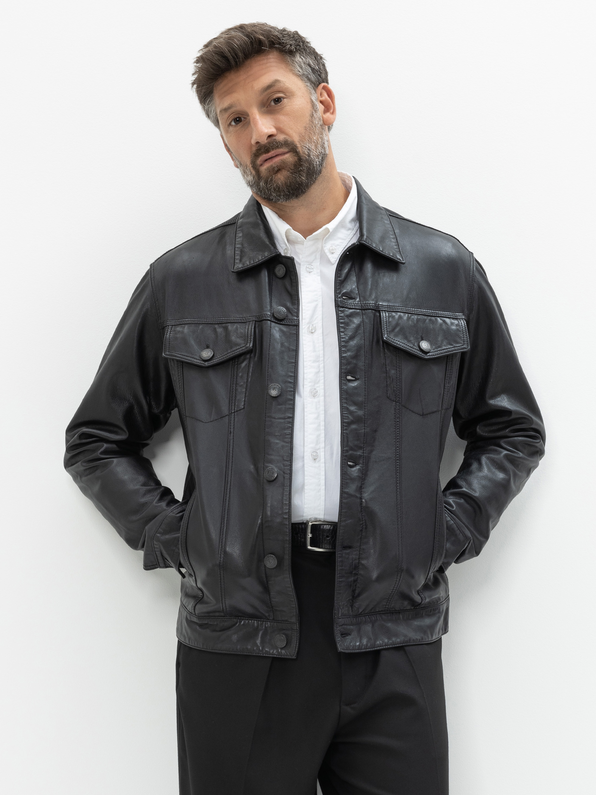 Ein Mann mit kurzen braunen Haaren und Bart trägt eine schwarze Lederjacke über einem weißen Button-up-Hemd und einer schwarzen Hose. Er steht vor einem schlichten weißen Hintergrund und hat die Hände in den Taschen.