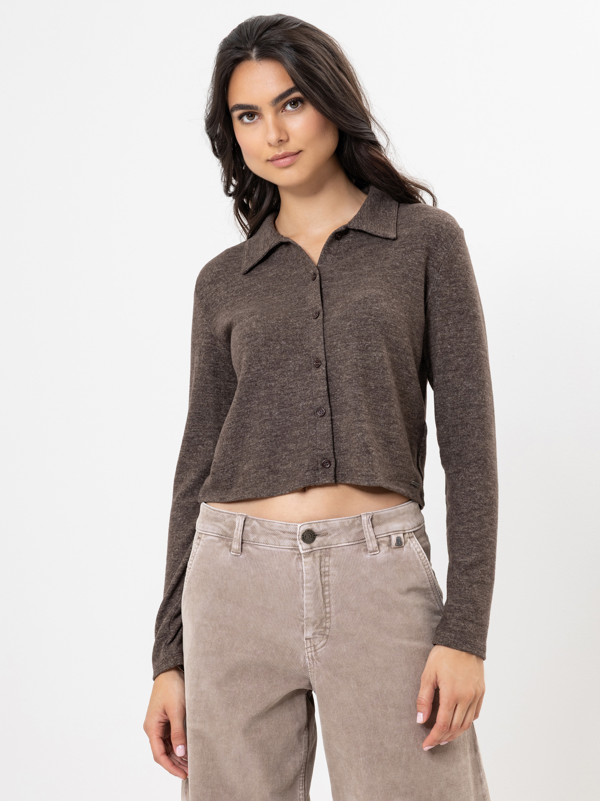 Eine Frau mit langen dunklen Haaren trägt ein braunes, langärmeliges, geknöpftes Oberteil und eine hellbeige Hose mit hoher Taille vor einem schlichten weißen Hintergrund.