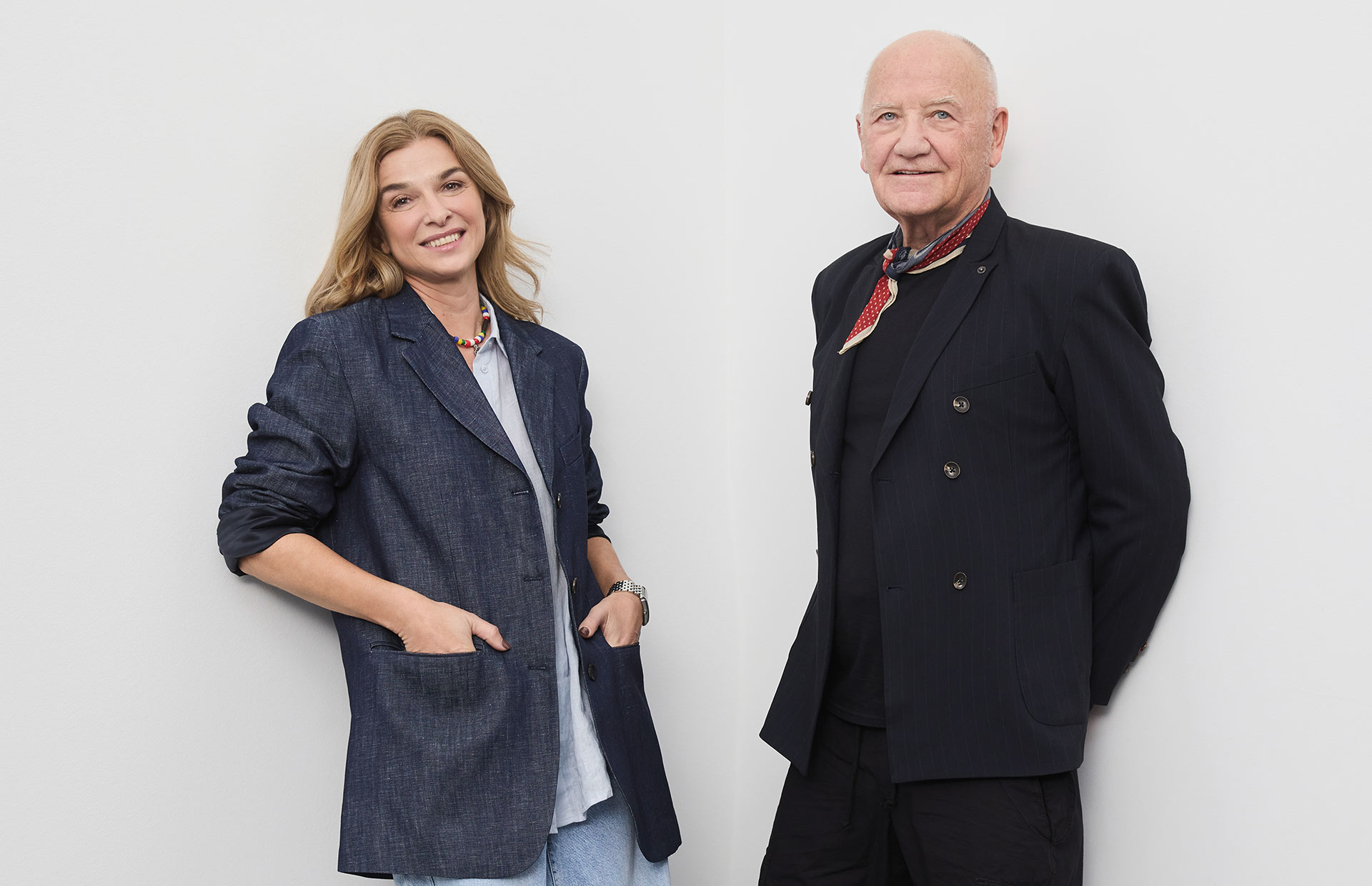 Eine Frau in einem blauen Blazer und ein Mann in einem dunklen Anzug stehen Seite an Seite vor einer schlichten weißen Wand und lächeln in die Kamera.