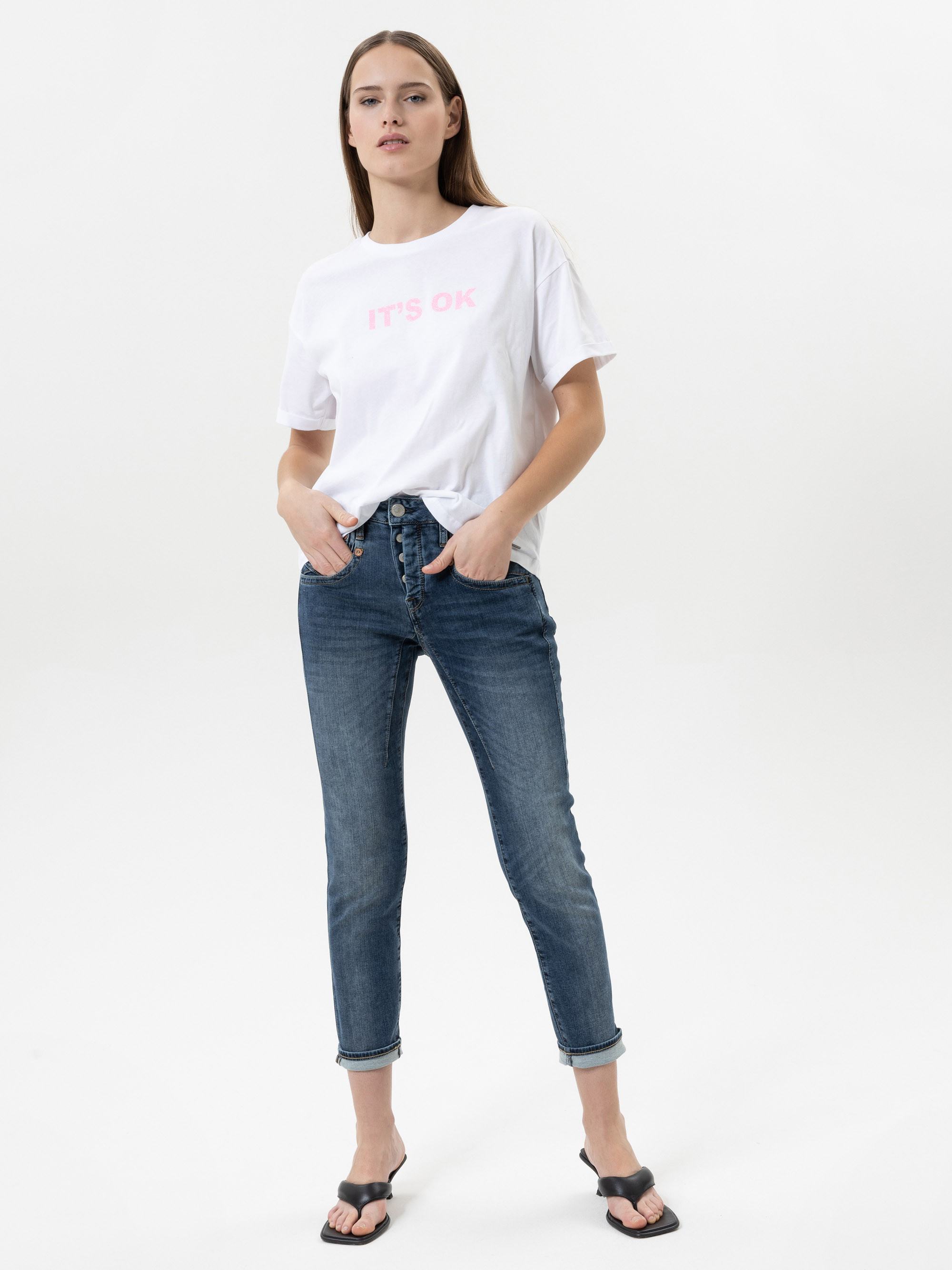 Eine Frau steht vor einem schlichten weißen Hintergrund. Sie trägt ein weißes T-Shirt mit der rosa Aufschrift ITS OK, blaue Jeans mit Bündchen und schwarze Sandalen. Sie hat glattes braunes Haar und ihre Hände stecken in den Taschen.