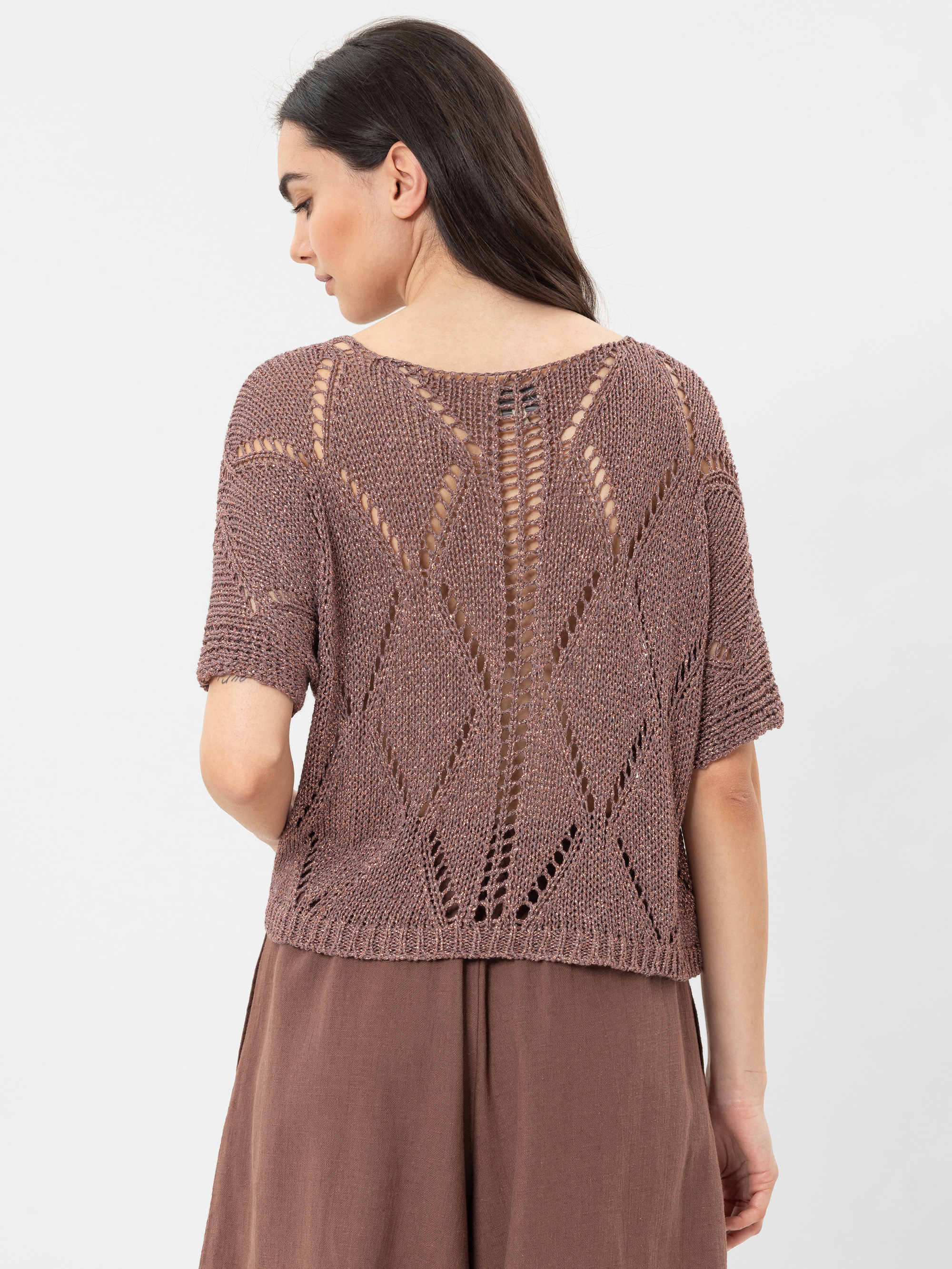 Eine Frau mit langen dunklen Haaren trägt ein locker sitzendes, kurzärmeliges, lilafarbenes Strickoberteil mit einem geometrischen, durchbrochenen Muster auf dem Rücken, kombiniert mit einer braunen, hoch sitzenden Hose. Sie schaut von der Kamera weg.