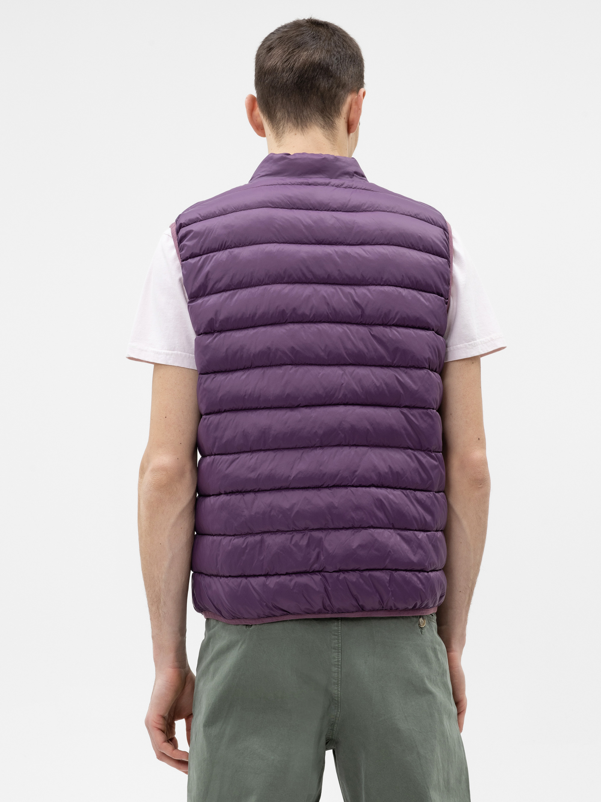 Eine Person wird von hinten gezeigt, die eine lila Pufferweste über einem kurzärmeligen weißen Hemd und eine grüne Hose trägt und vor einem einfarbig weißen Hintergrund steht.
