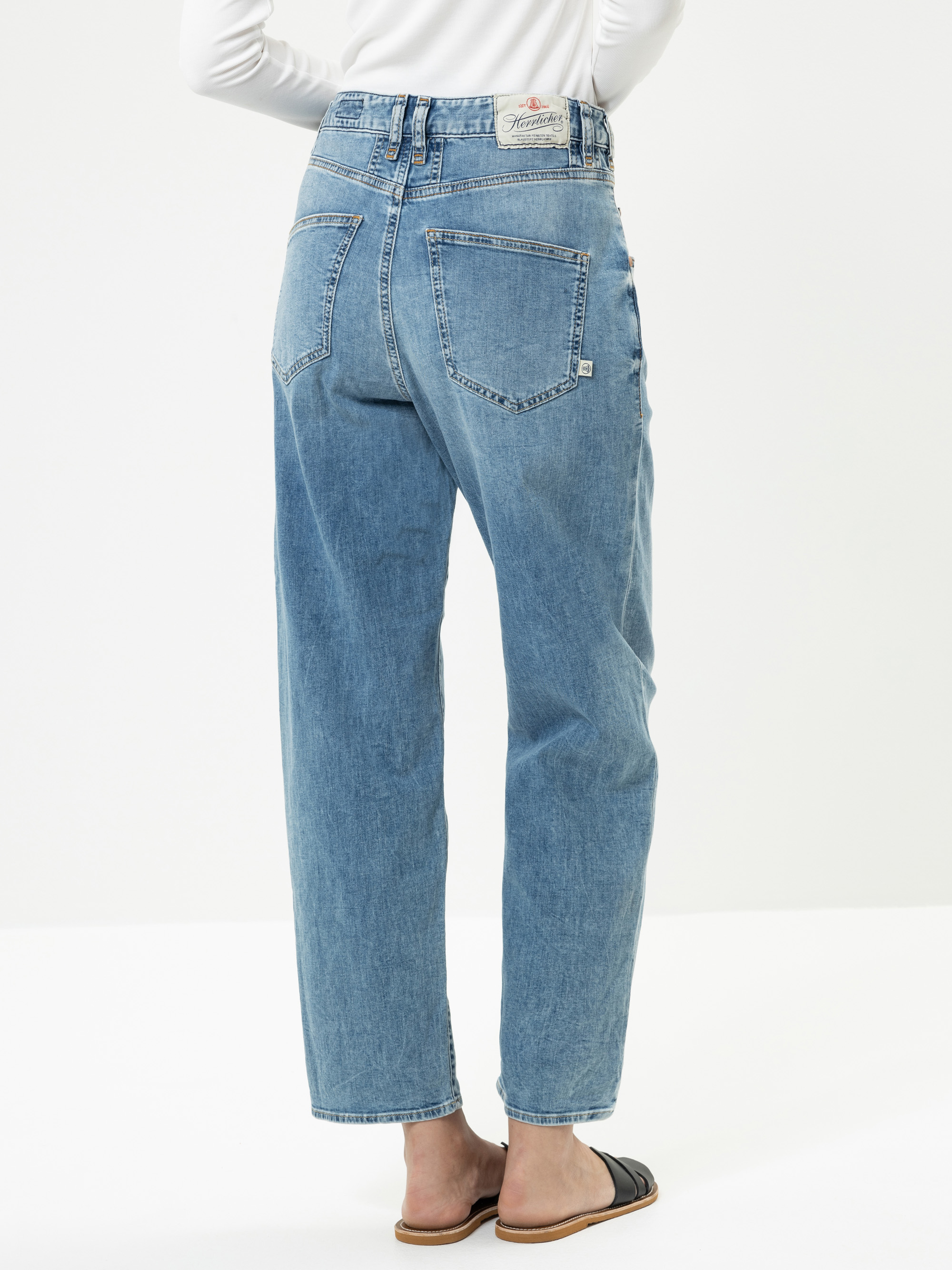 Eine Person, die hochgeschnittene, locker sitzende Bluejeans und schwarze Sandalen trägt, ist von hinten zu sehen und steht vor einem schlichten weißen Hintergrund.