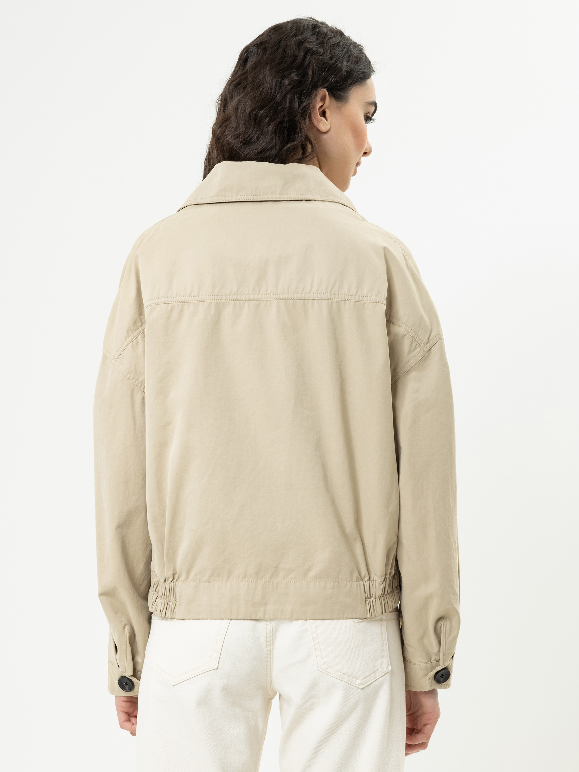 Eine Frau mit gewelltem dunklem Haar ist von hinten zu sehen. Sie trägt eine locker sitzende beigefarbene Jacke mit geknöpften Manschetten und eine weiße Hose vor einem schlichten weißen Hintergrund.