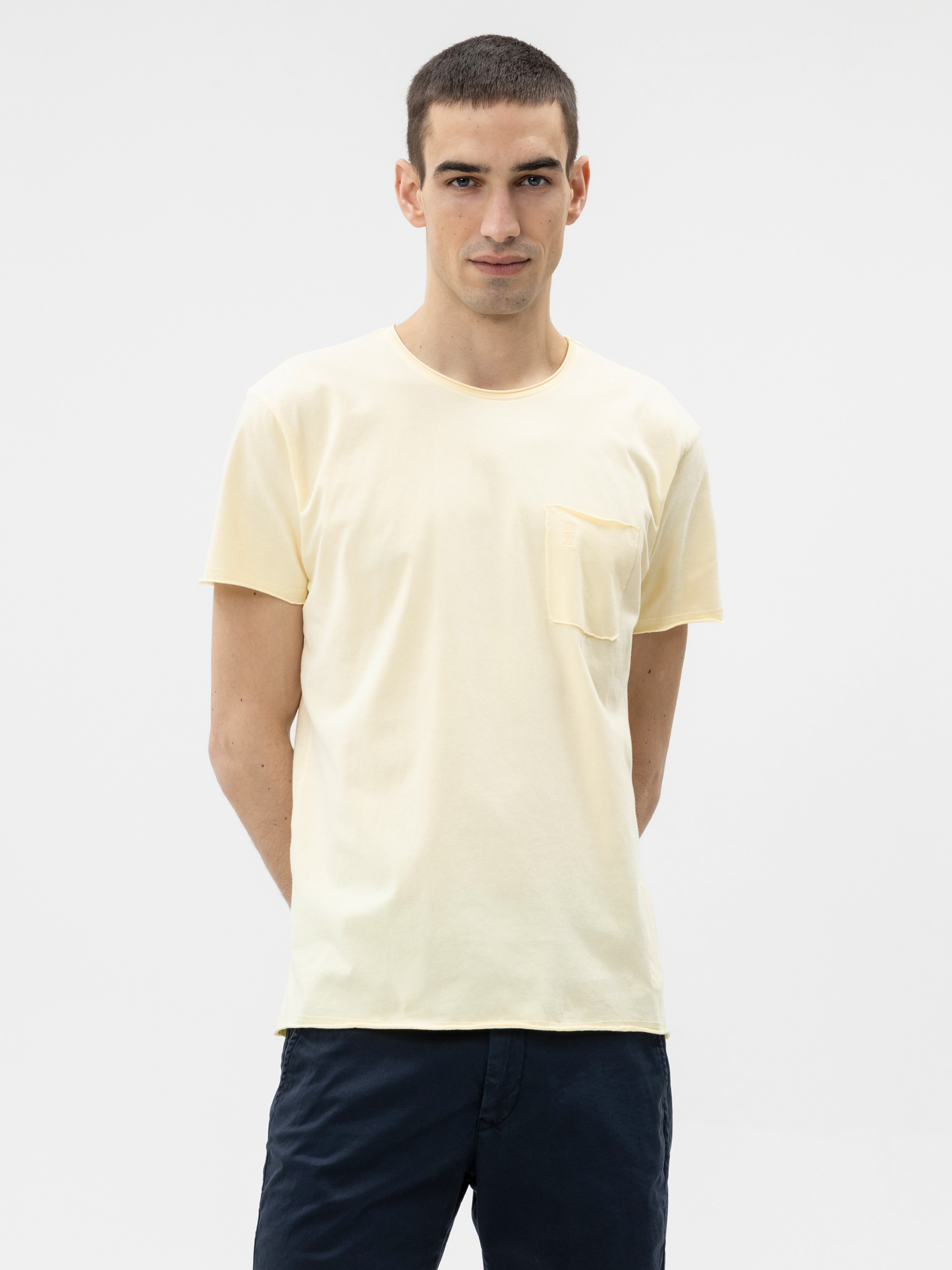 Ein Mann mit kurzen dunklen Haaren steht vor einem schlichten hellen Hintergrund. Er trägt ein hellgelbes T-Shirt mit einer Brusttasche und dunkle Shorts. Er verschränkt die Arme hinter dem Rücken und hat einen neutralen Gesichtsausdruck.