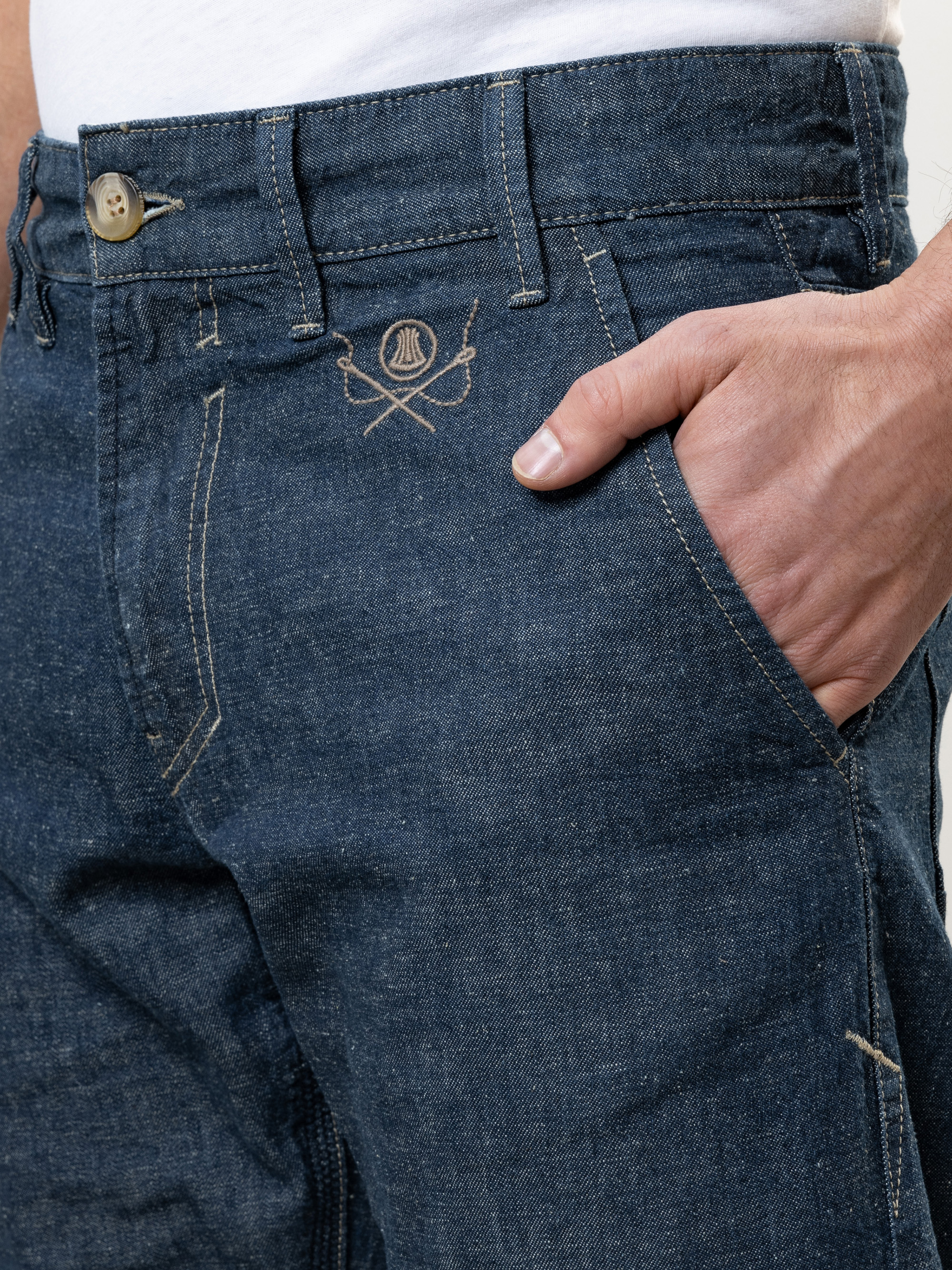 Eine Person, die eine blaue Jeans und ein weißes Hemd trägt, hat die rechte Hand in der Vordertasche. Die Jeans hat ein kleines gesticktes Logo mit gekreuzten Paddeln unterhalb des Hosenbundes.