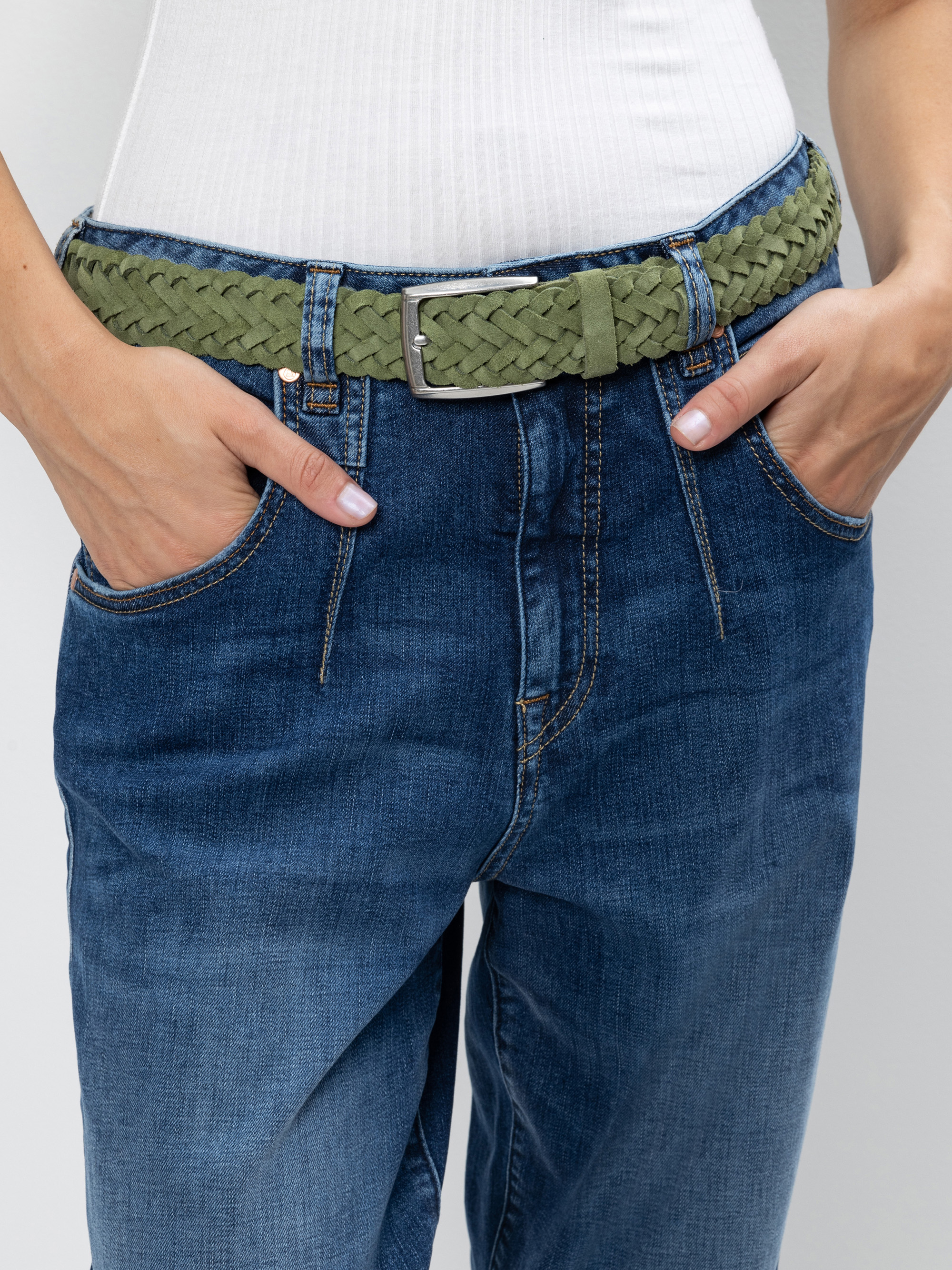 Eine Person, die blaue Jeans, ein weißes geripptes Oberteil und einen olivgrünen Flechtgürtel trägt, steht mit den Händen in den Vordertaschen vor einem neutralen Hintergrund.