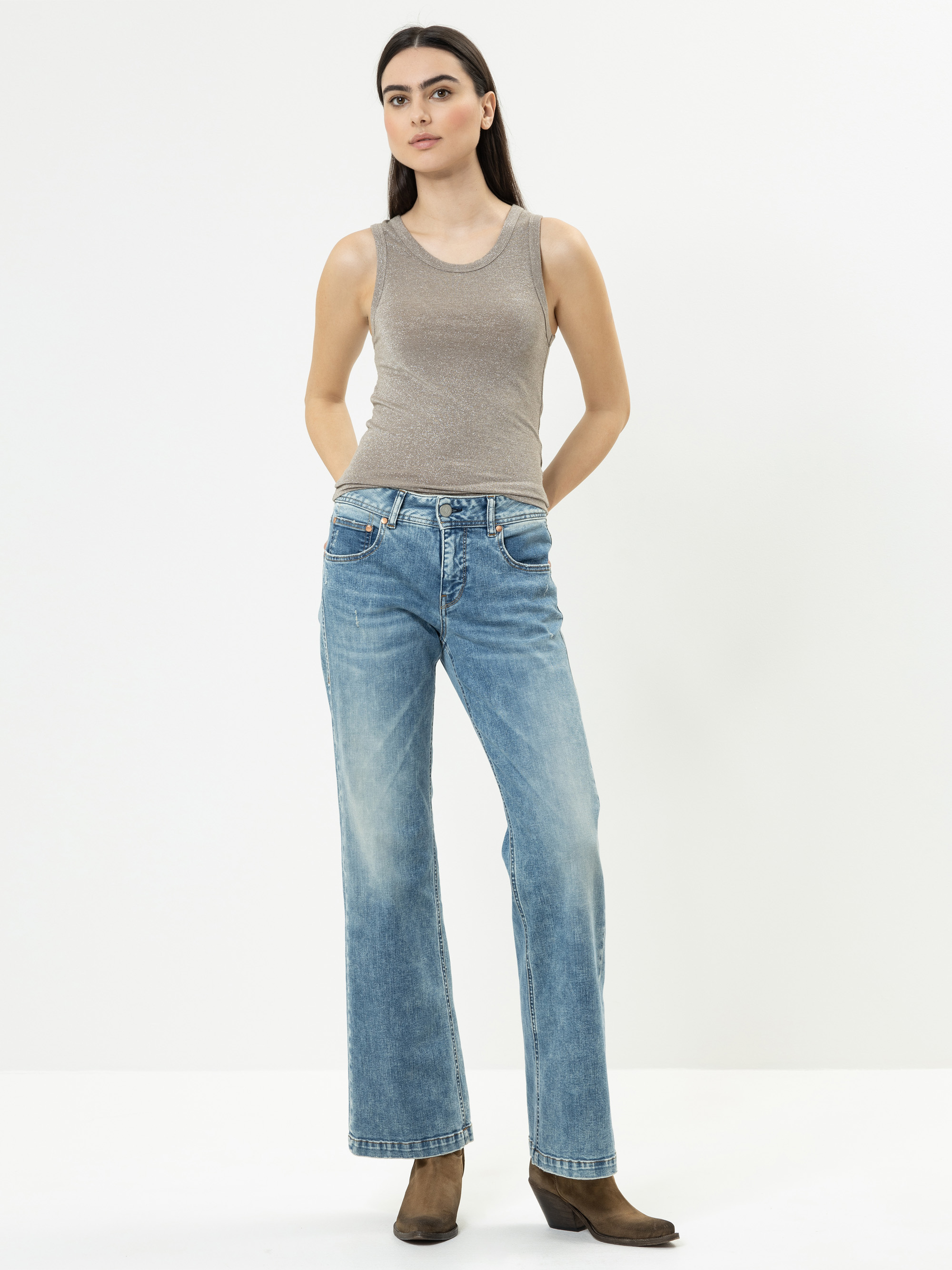 Eine Frau steht vor einem schlichten weißen Hintergrund und trägt ein ärmelloses beiges Oberteil, blaue Jeans mit geradem Bein und braune Stiefeletten. Sie hat langes dunkles Haar und schaut leicht zur Seite, die Hände in den Vordertaschen.