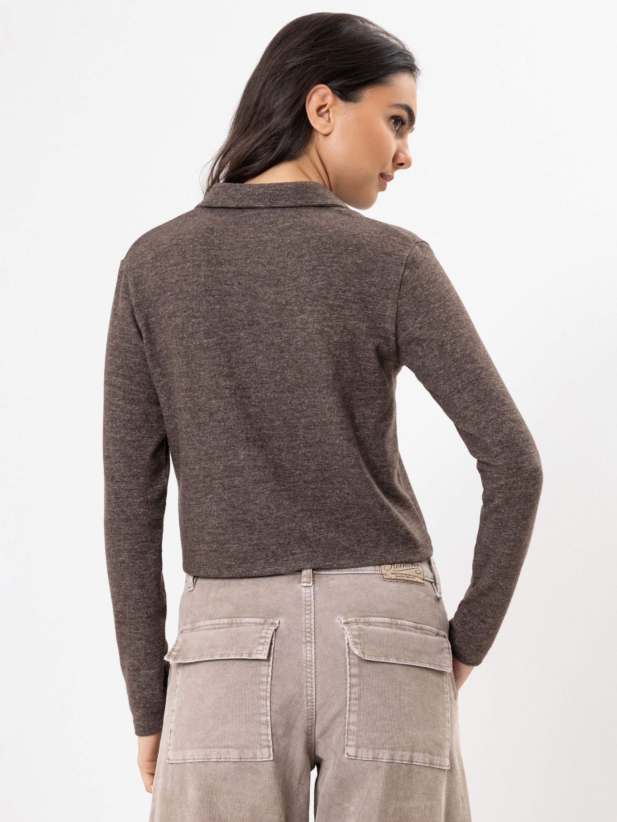 Eine Frau mit langen dunklen Haaren trägt ein braunes langärmeliges Oberteil und eine hellgraue Hose mit großen Gesäßtaschen und steht mit dem Rücken zur Kamera vor einem schlichten weißen Hintergrund.