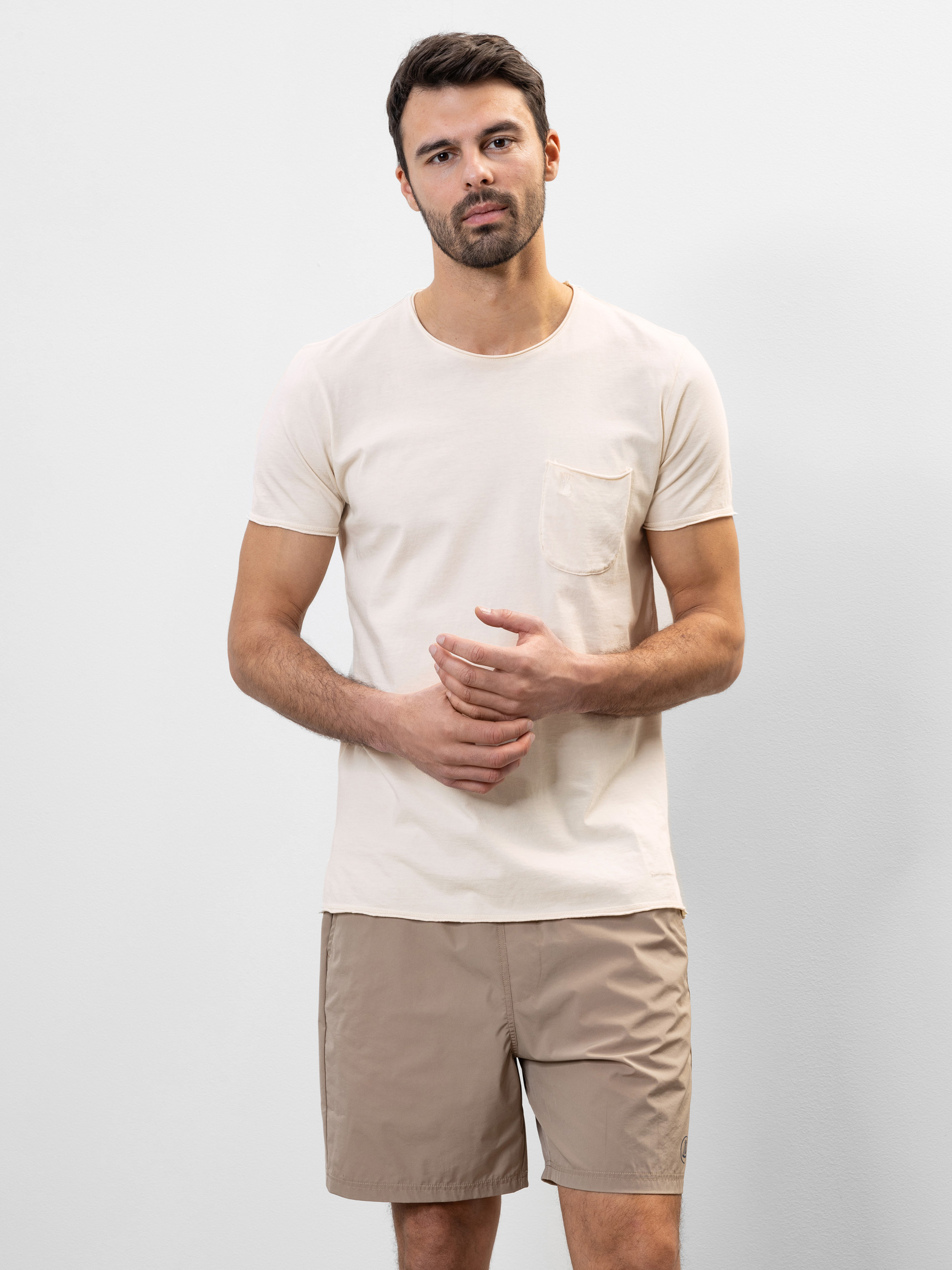 Ein Mann mit kurzen dunklen Haaren und Bart trägt ein hellbeiges T-Shirt mit einer Brusttasche und hellbraune Shorts. Er steht vor einem schlichten hellgrauen Hintergrund und blickt mit neutralem Blick in die Kamera.