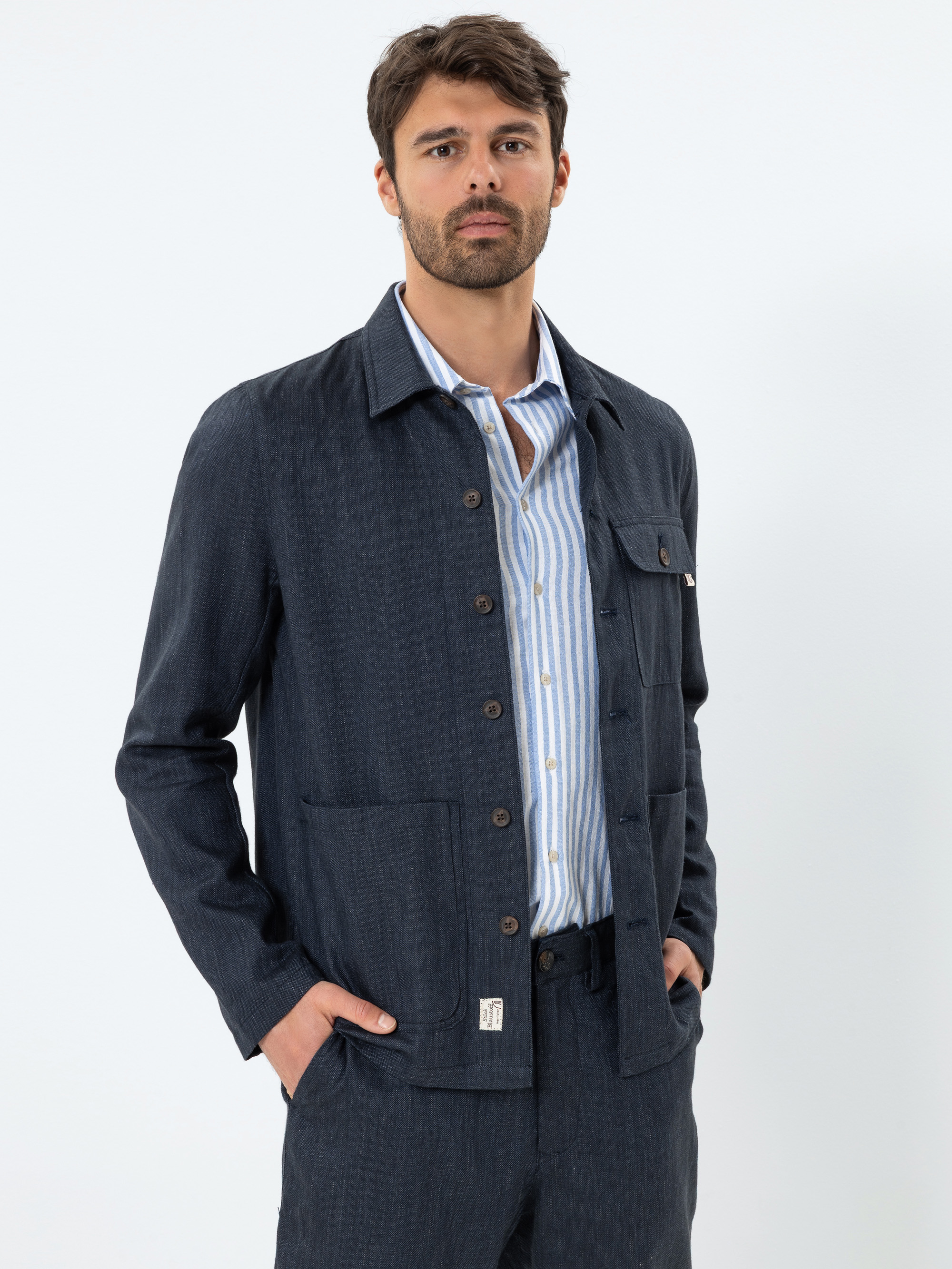 Ein Mann mit braunem Haar und Bart trägt eine dunkelblaue Jacke über einem hellblau-weiß gestreiften Hemd und posiert mit den Händen in den Taschen vor einem schlichten weißen Hintergrund.