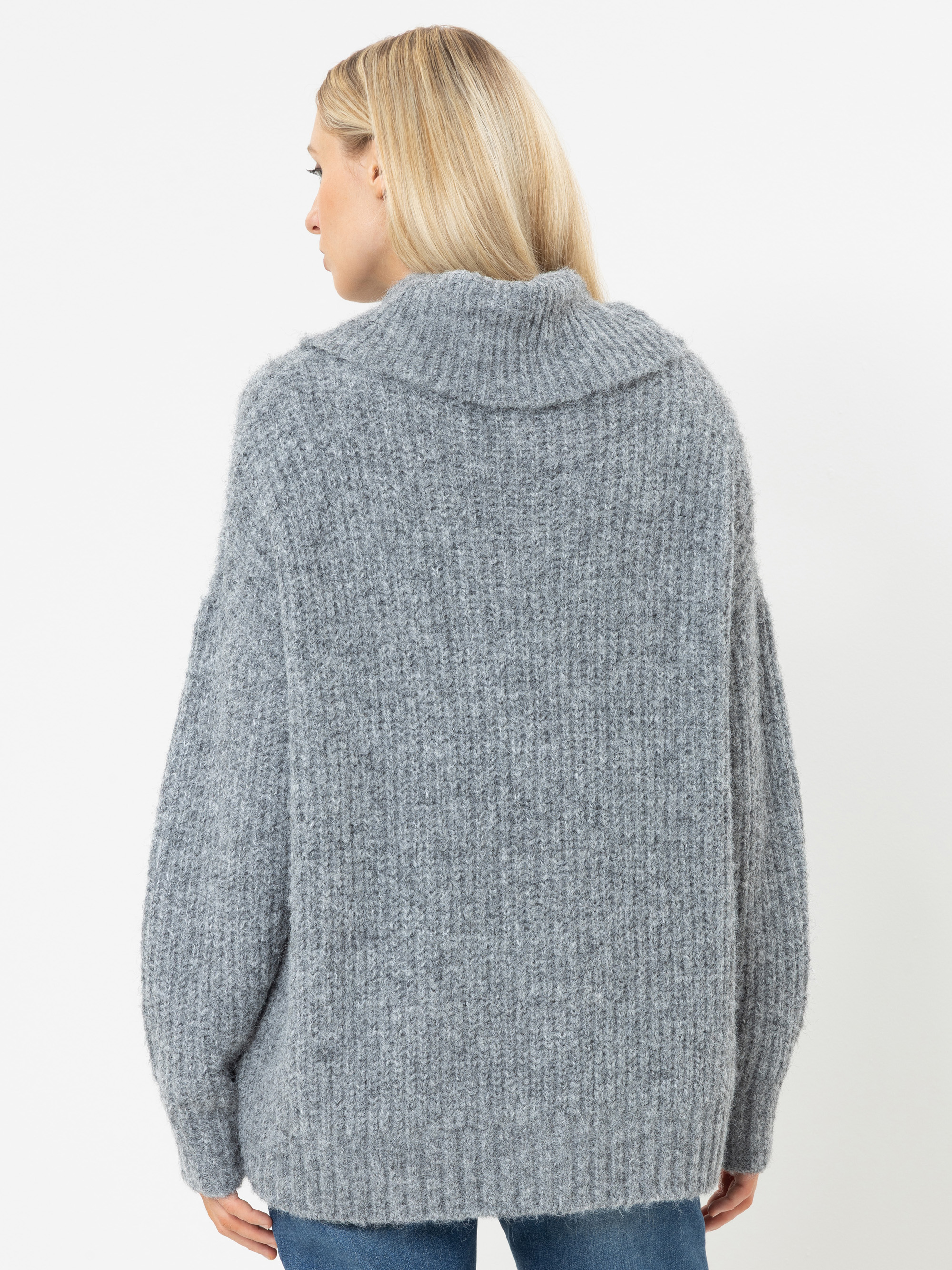 Eine Person mit langen blonden Haaren, von hinten gesehen, trägt einen grob gestrickten, hellgrauen Pullover mit Rippenmuster und hohem Kragen. Außerdem trägt sie eine blaue Jeans und steht vor einem schlichten weißen Hintergrund.