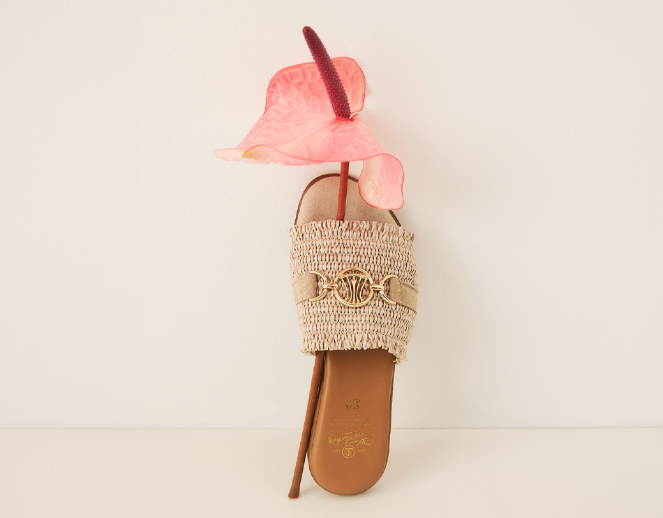 Eine gewebte beigefarbene Sandale mit einem goldenen Schmuckemblem ist aufrecht platziert, und eine rosa Anthurienblüte ist durch die Sandale hindurch positioniert, wobei der Stiel unten herausragt. Der Hintergrund ist einfarbig cremefarben.