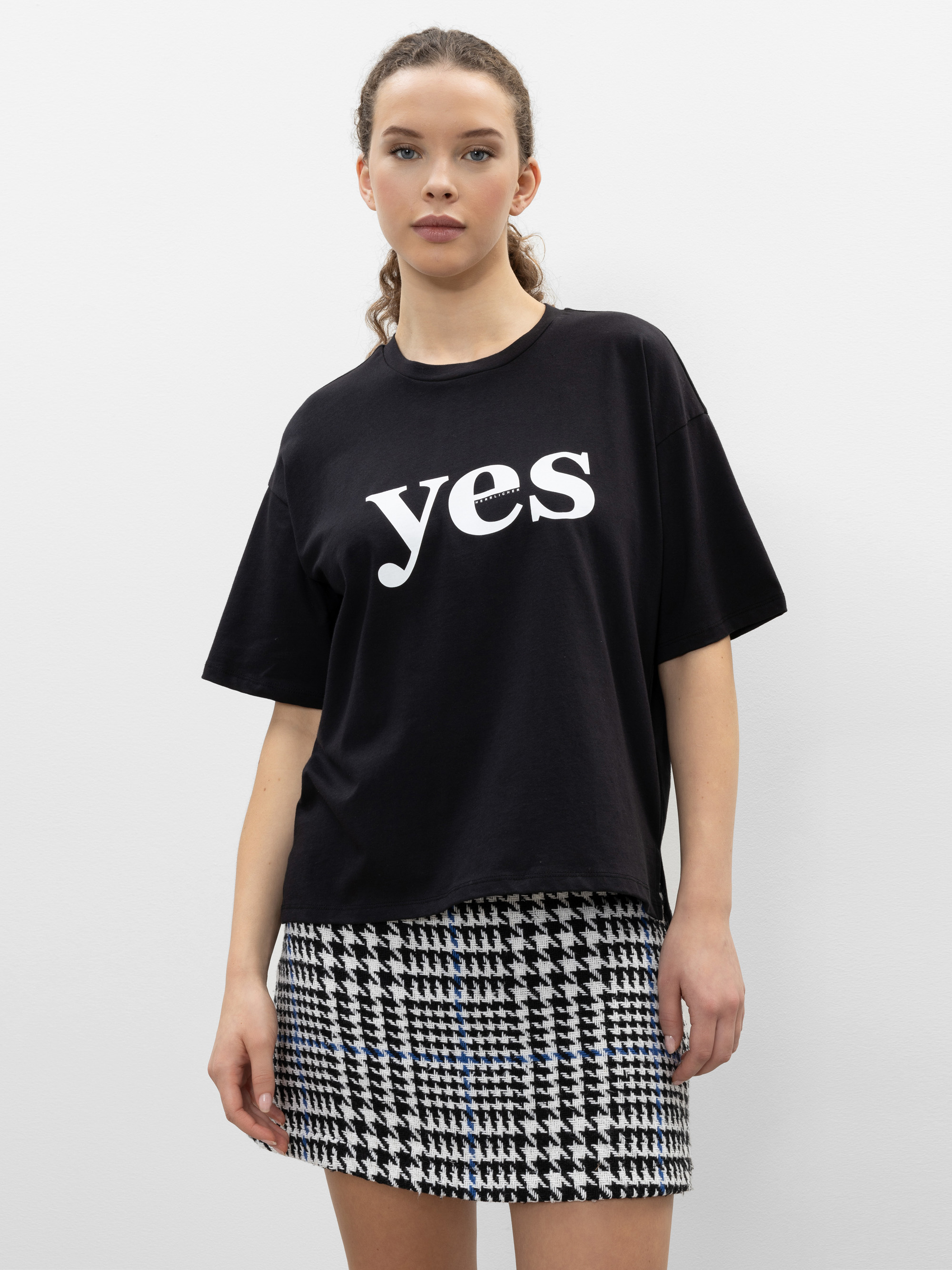 Eine Frau steht vor einem schlichten Hintergrund und trägt ein schwarzes T-Shirt mit dem Wort "Yes" in fetten weißen Buchstaben und einen schwarz-weißen Hahnentritt-Rock. Sie hat helle Haut, zurückgebundenes, gewelltes Haar und einen neutralen Gesicht