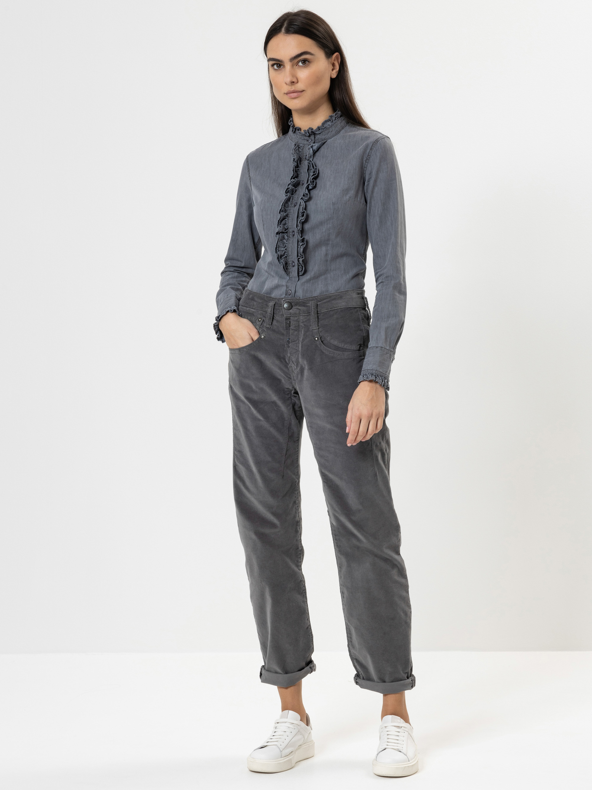 Eine Frau steht vor einem schlichten weißen Hintergrund und trägt eine langärmelige, gerüschte graue Bluse, eine graue Hose mit hoher Taille und aufgerollten Manschetten sowie weiße Turnschuhe. Sie hat langes dunkles Haar und einen neutralen Gesichts