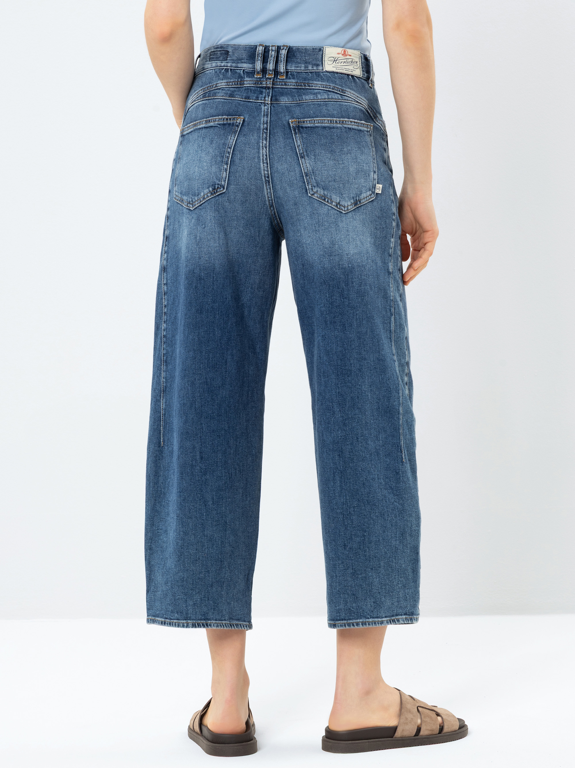 Die Person trägt eine blaue Jeans mit weitem Bein und verblasster Waschung, gepaart mit braunen Sandalen. Die Abbildung zeigt die Rückenansicht vor einem schlichten weißen Hintergrund.