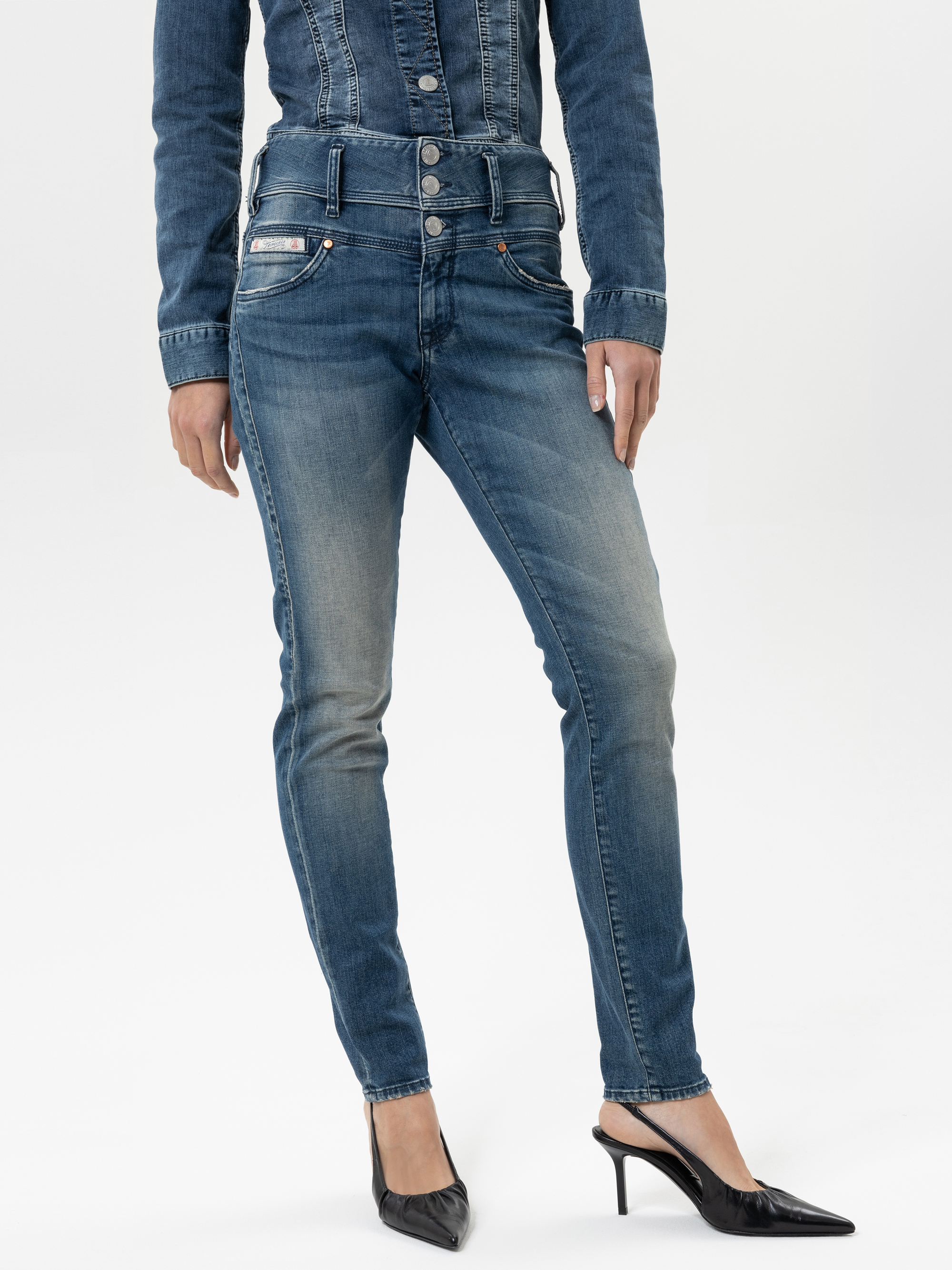 Eine Person, die hochtaillierte blaue Skinny-Jeans mit mehreren Knöpfen und eine dazu passende Jeansjacke trägt, steht mit schwarzen Schuhen mit spitzen Absätzen vor einem schlichten weißen Hintergrund.