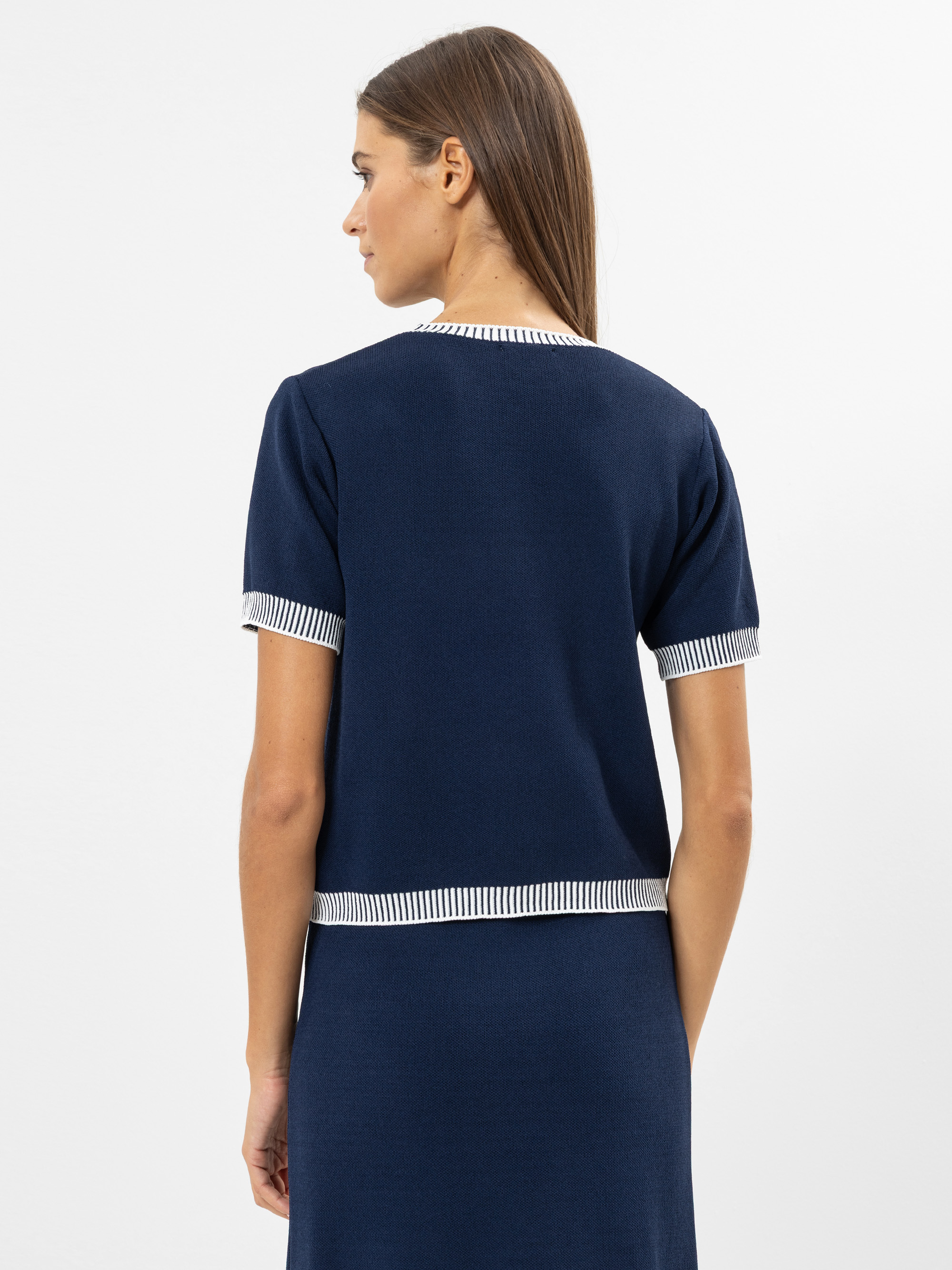 Eine Frau mit glattem braunem Haar ist von hinten zu sehen. Sie trägt ein marineblaues kurzärmeliges Oberteil mit weiß und marineblau gestreiften Abschlüssen an Ärmeln, Ausschnitt und Saum sowie einen passenden Rock.
