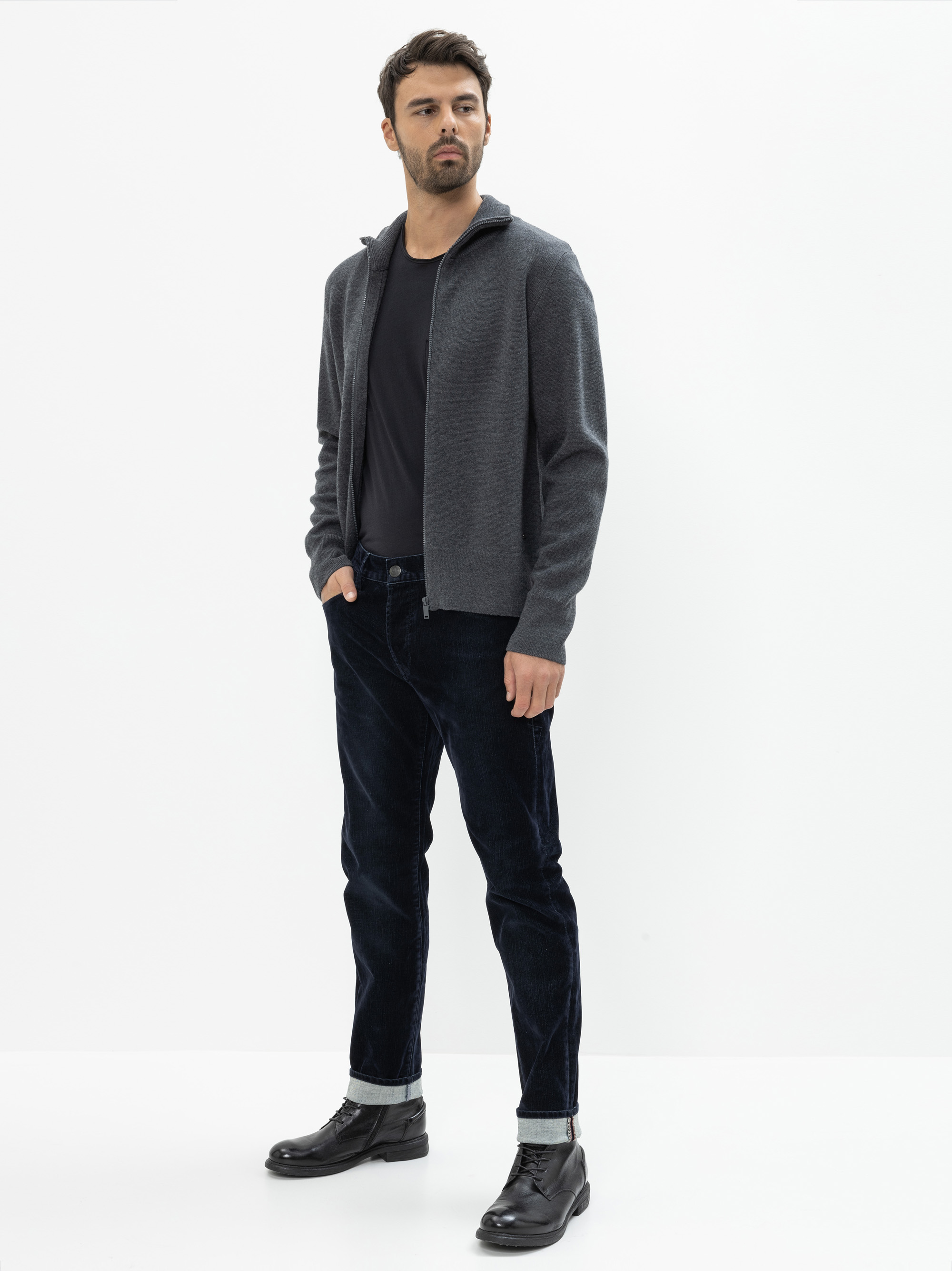 Ein Mann steht vor einem schlichten weißen Hintergrund und trägt eine graue Strickjacke mit Reißverschluss, ein schwarzes T-Shirt, dunkle Jeans mit Bündchen und schwarze Stiefel. Er hat die Hände in den Taschen und blickt leicht zur Seite.