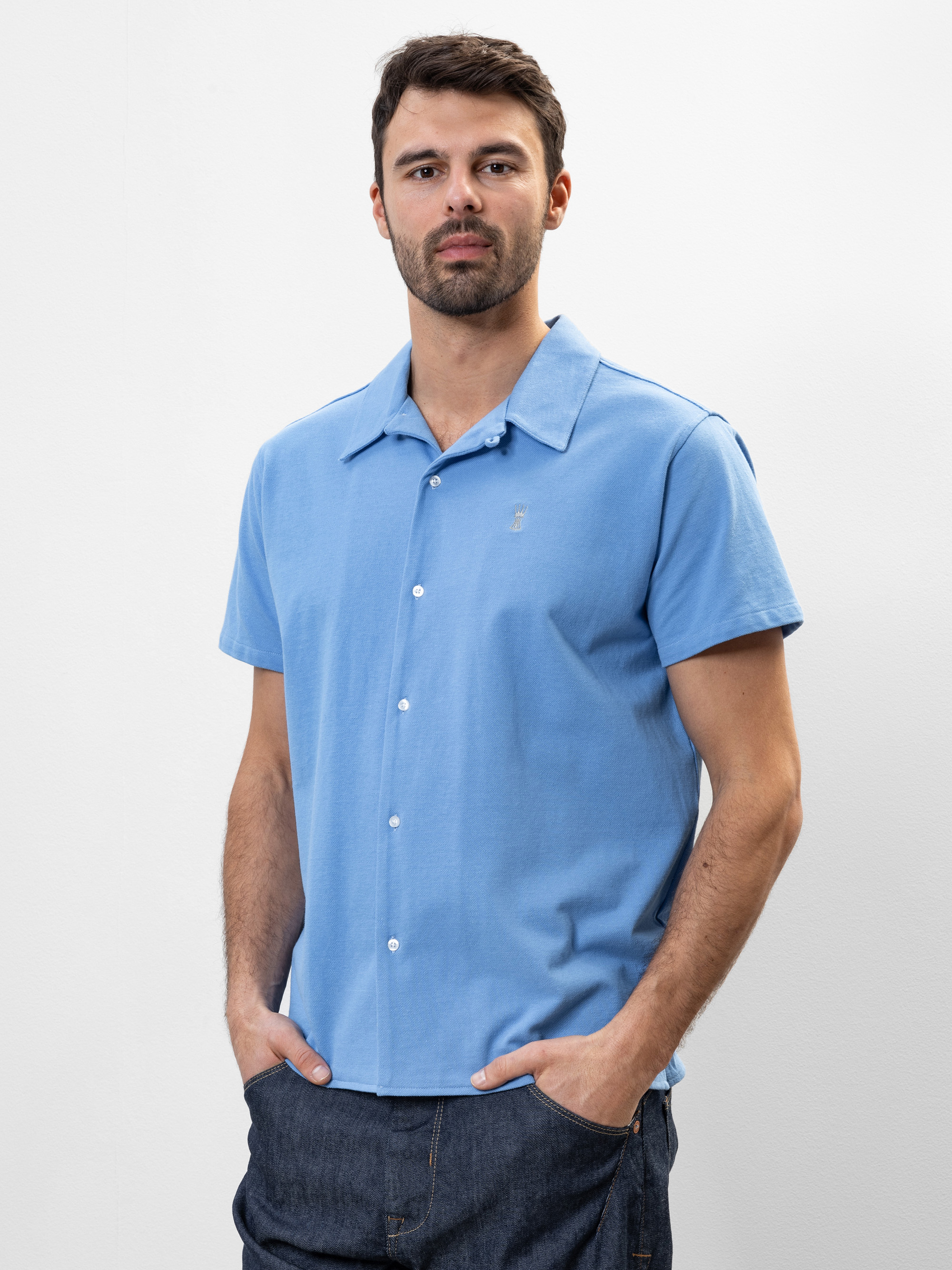 Ein Mann mit kurzen braunen Haaren und einem Bart trägt ein hellblaues, kurzärmeliges Button-up-Hemd und dunkle Jeans. Er steht vor einem schlichten weißen Hintergrund und hat die Hände in den Taschen.