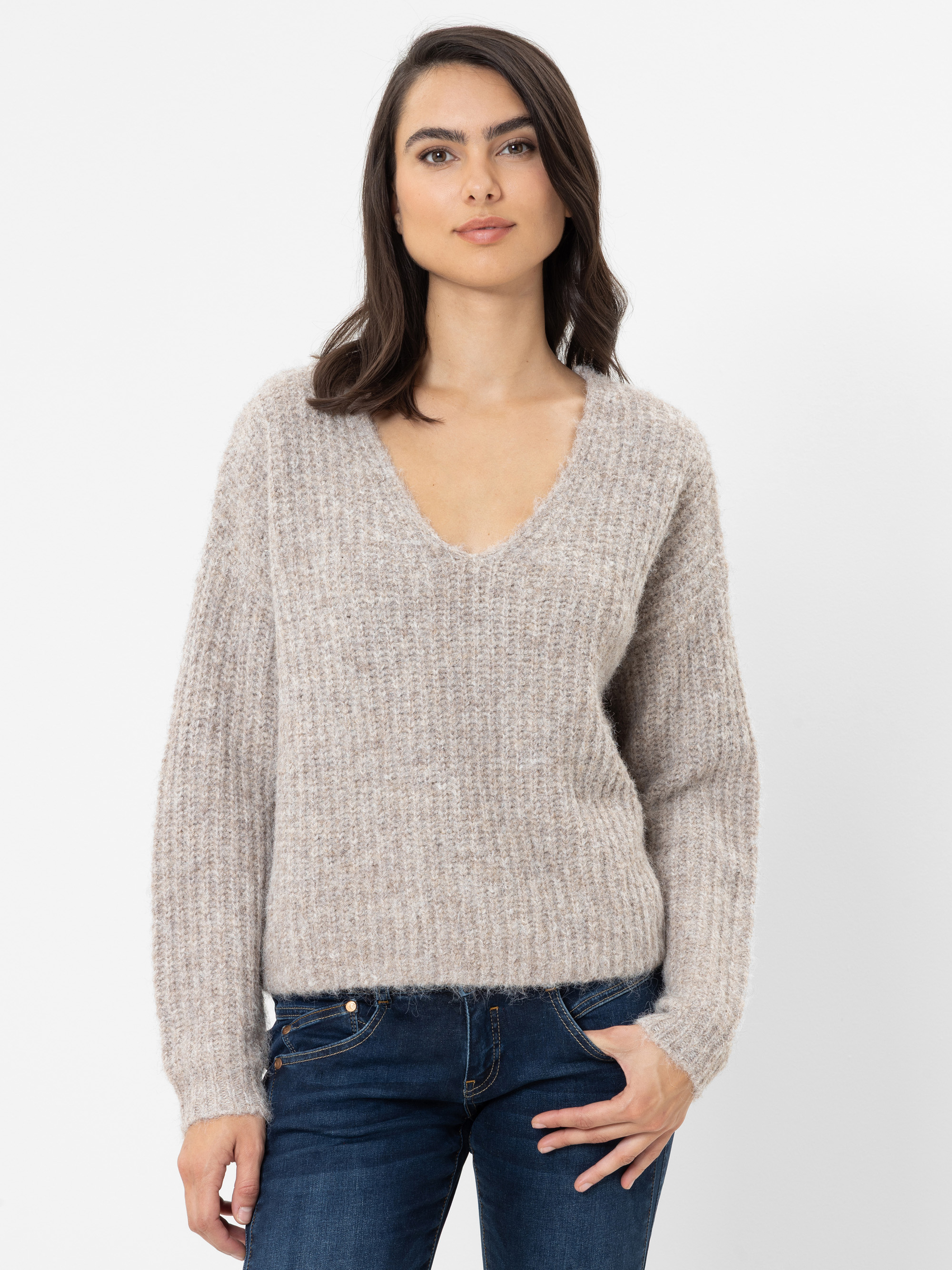 Eine Frau mit glattem braunem Haar, die einen beigen, strukturierten Pullover mit V-Ausschnitt und blaue Jeans trägt, steht vor einem schlichten weißen Hintergrund mit entspanntem Gesichtsausdruck und einer Hand in der Hosentasche.