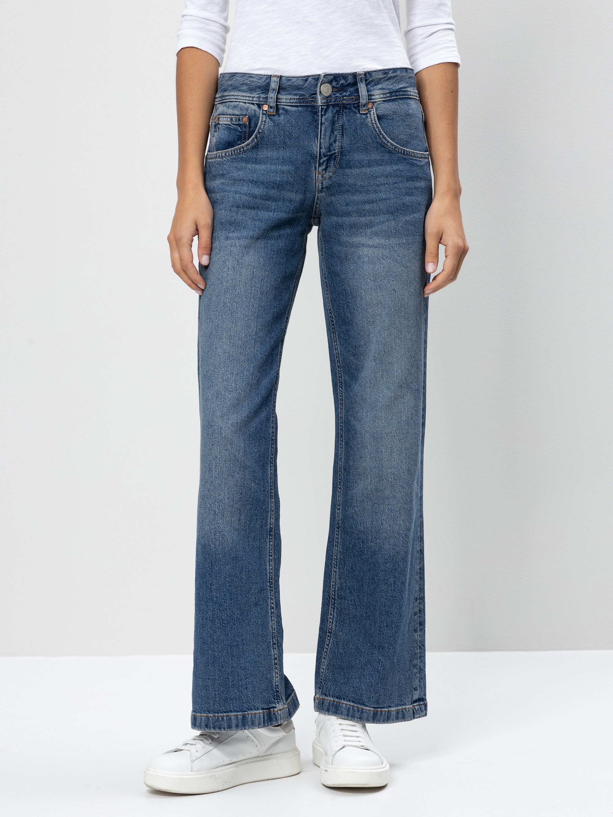 Eine Person, die ein weißes langärmeliges Hemd, eine blaue Jeans mit geradem Bein und weiße Turnschuhe trägt, steht vor einem schlichten hellgrauen Hintergrund. Nur der Körper von den Schultern bis zu den Füßen ist sichtbar.