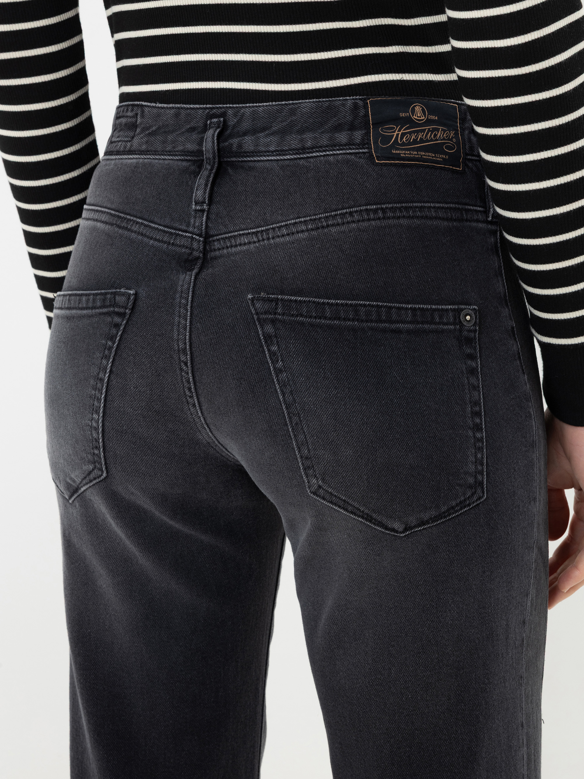 Eine Person, die schwarze Jeans und ein schwarz-weiß gestreiftes Oberteil trägt, von hinten gesehen. Die Jeans hat am Bund ein braunes Aufnäher-Etikett, auf dem der Markenname „Herrlicher“ zu sehen ist.