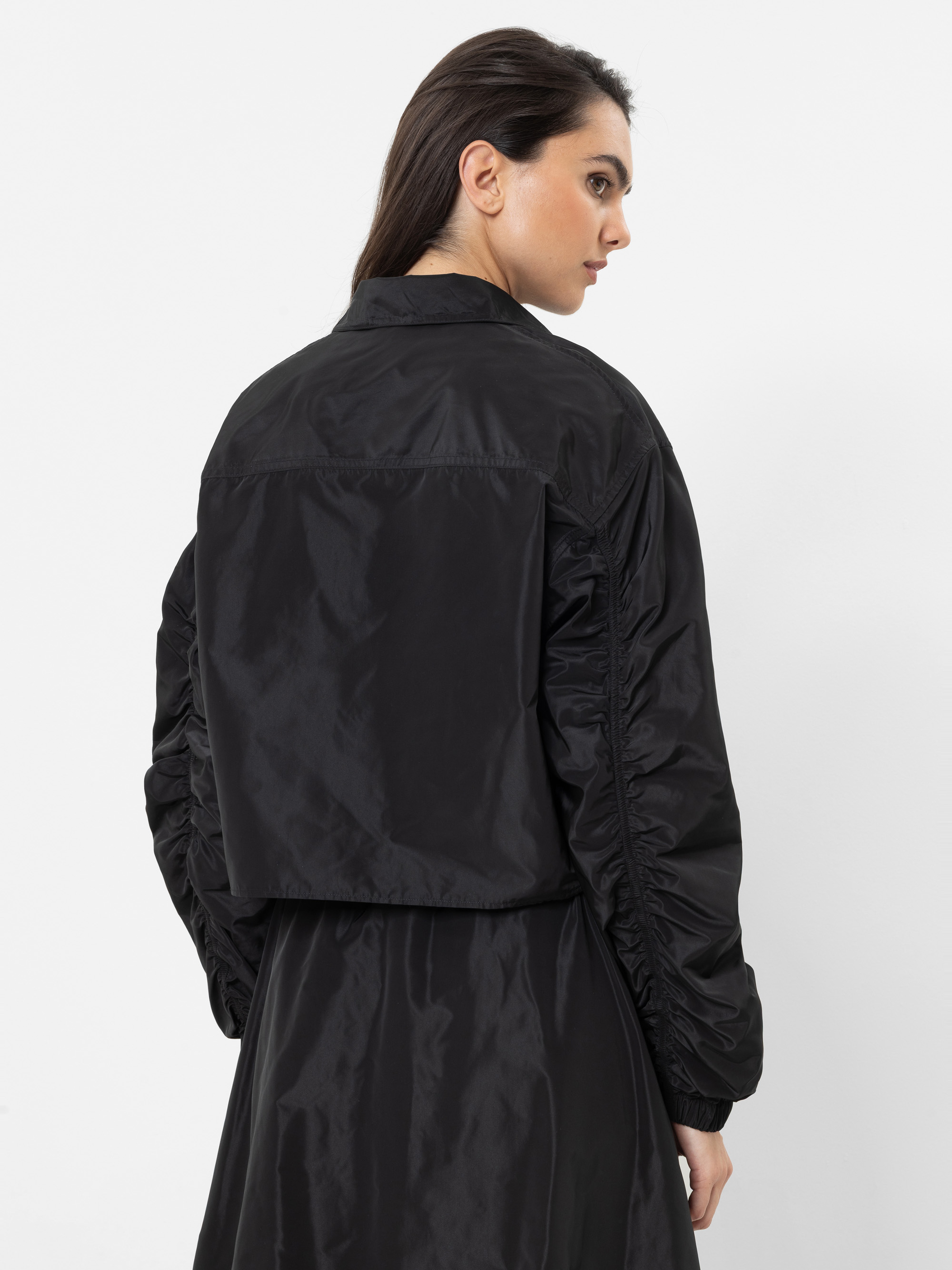 Eine Frau mit langen dunklen Haaren, die eine glänzende schwarze kurze Jacke über einem passenden schwarzen Kleid trägt, steht vor einem schlichten weißen Hintergrund und blickt über ihre linke Schulter.