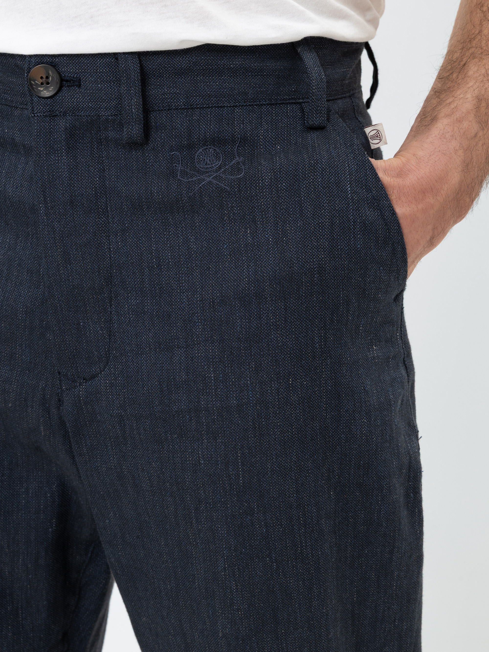 Eine Person, die eine dunkelblaue strukturierte Hose und ein weißes Hemd trägt und die rechte Hand in der Tasche hat. Die Hose hat einen Knopfverschluss und ein kleines aufgenähtes Emblem in der Nähe der Tasche.