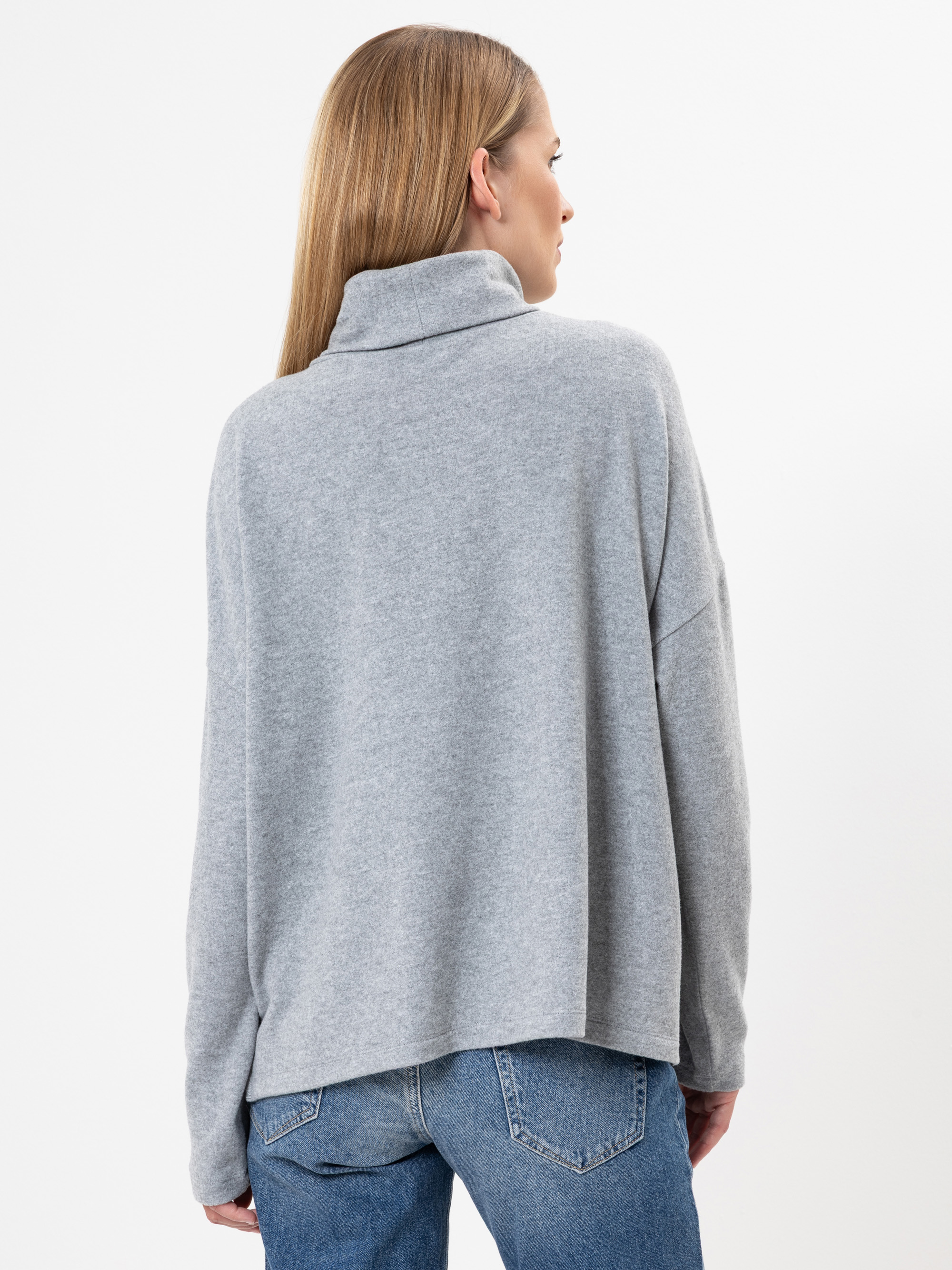 Eine Frau mit glattem, hellbraunem Haar trägt einen locker sitzenden, hellgrauen Rollkragenpullover und blaue Jeans. Sie steht mit dem Rücken zur Kamera vor einem schlichten weißen Hintergrund.
