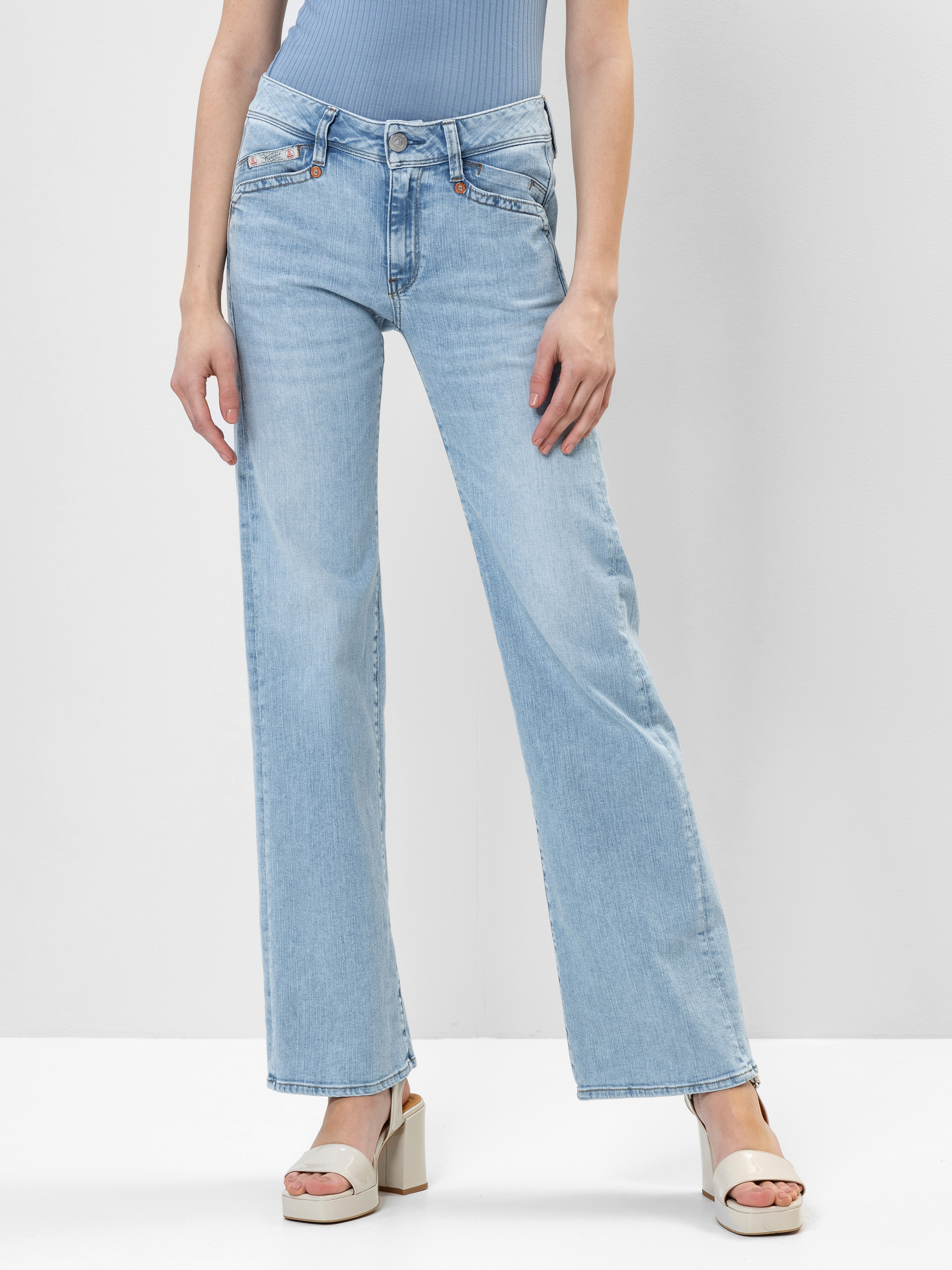 Eine Person in hellblauen, weit geschnittenen Jeans und Plateausandalen steht vor einem schlichten weißen Hintergrund. Ihre Arme liegen entspannt an der Seite.