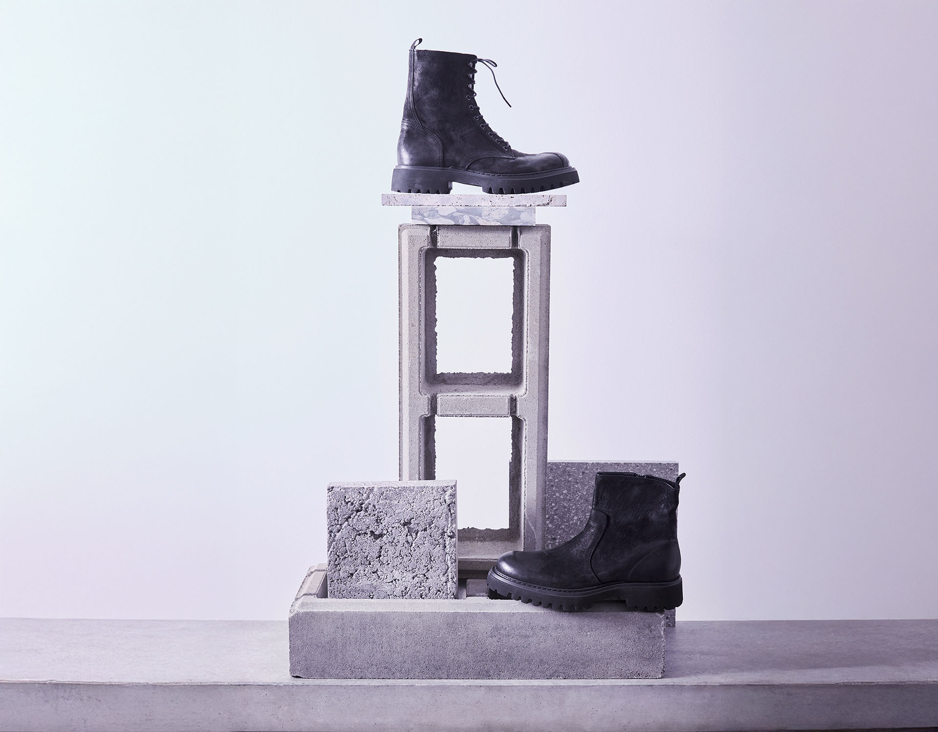 Zwei schwarze Stiefel sind auf gestapelten Betonblöcken und strukturierten Steinfliesen vor einem hellvioletten Hintergrund ausgestellt, wobei ein Stiefel oben auf den Blöcken und der andere unten auf der Oberfläche liegt.