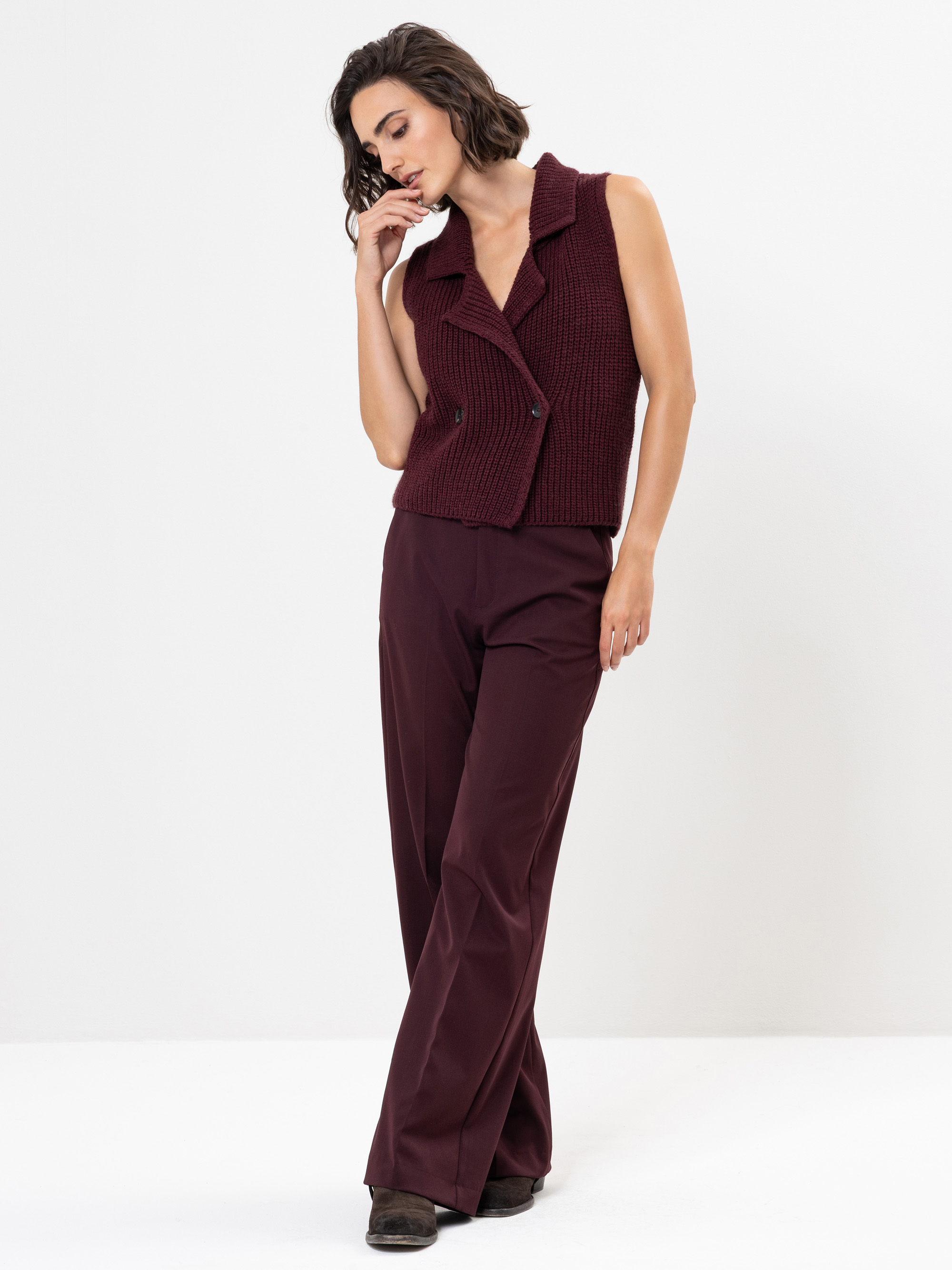 Eine Frau in einer ärmellosen, dunkelroten Strickweste und einer dazu passenden Hose mit weitem Bein steht vor einem weißen Hintergrund und blickt nach unten, wobei eine Hand sanft ihr Gesicht berührt.