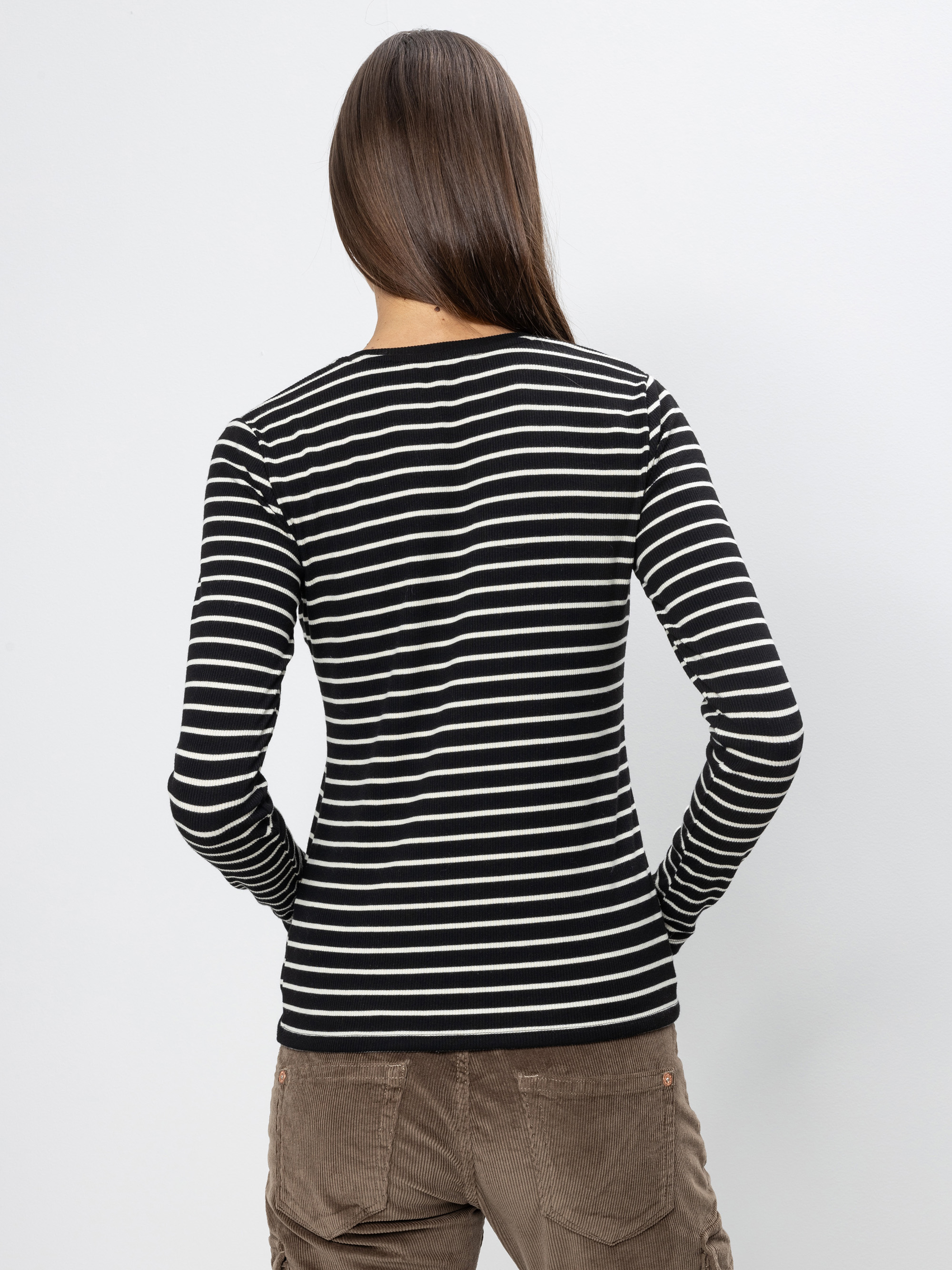 Gezeigt wird eine Person mit langen braunen Haaren von hinten, die ein schwarz-weiß gestreiftes langärmeliges Hemd und eine braune Hose vor einem einfarbigen hellgrauen Hintergrund trägt.
