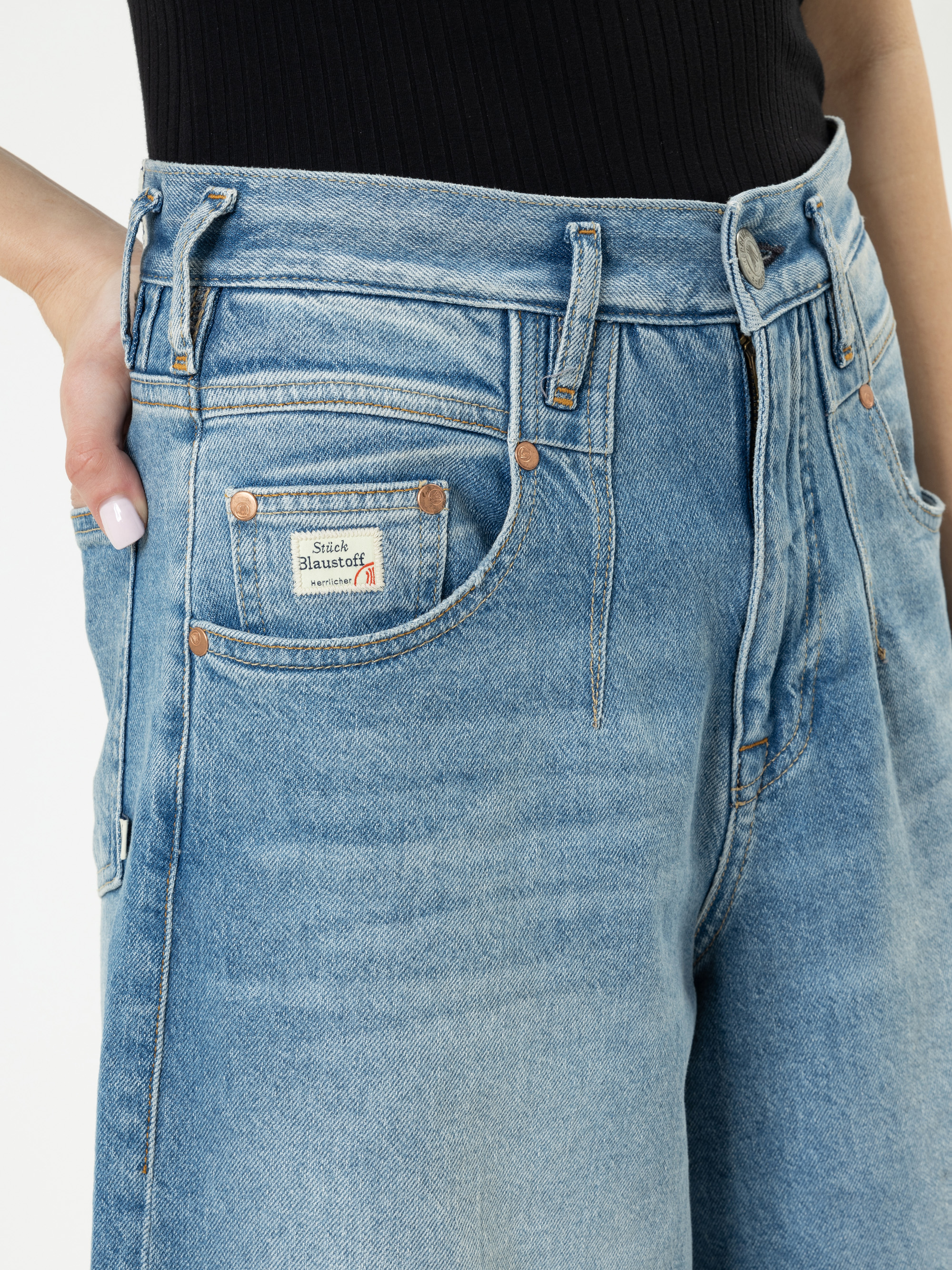 Eine Person, die eine hellblaue Jeans mit hoher Taille und ein schwarzes geripptes Oberteil trägt, hat die Hand in die Vordertasche gesteckt und zeigt ein kleines Etikett auf der Tasche, auf dem Studio Bannister steht.